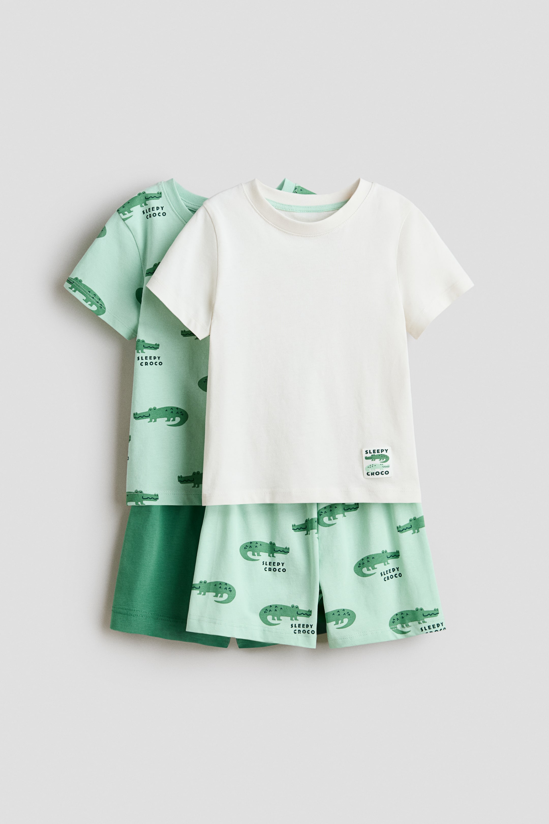 Ver imagem maior: Um conjunto de pijamas apresenta uma t-shirt branca lisa de manga curta com decote canelado e ombros ligeiramente descaídos, que é combinada com uns calções verdes claros exibindo um padrão repetido de crocodilos verdes e uma cintura elástica coberta. Um segundo conjunto de pijamas está parcialmente visível atrás, mostrando uma t-shirt verde clara com o mesmo padrão de crocodilos e uns calções verdes escuros lisos.