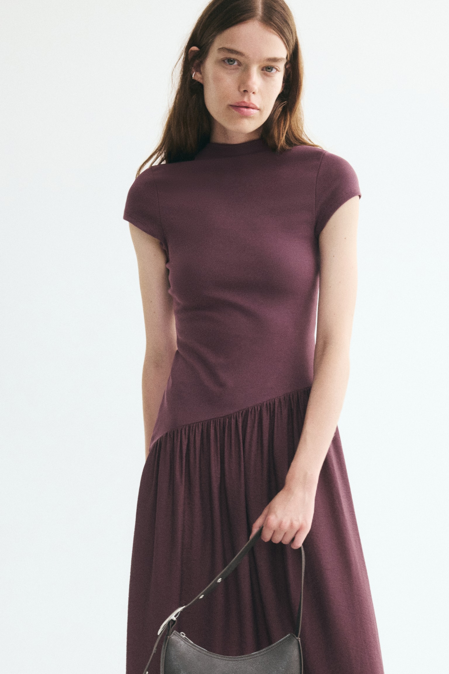 A maroon dress is hanging, facing front, featuring a mock neck and short sleeves. Its fitted bodice transitions into an asymmetrical, gathered seam, leading to a flowing, textured skirt that extends to a maxi length. - 1