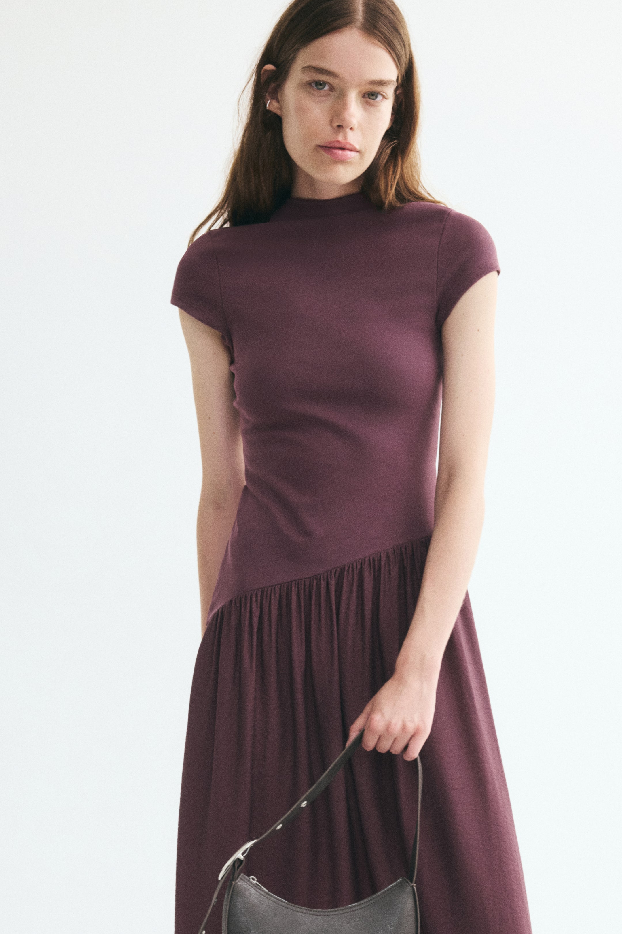 Agrandir l'image: Une robe longue prune foncé présente un corsage ajusté en maille côtelée, un col montant, des manches cap et une couture froncée asymétrique au niveau des hanches ; elle est portée par le mannequin. Le mannequin tient également un sac à bandoulière gris foncé par sa bandoulière.