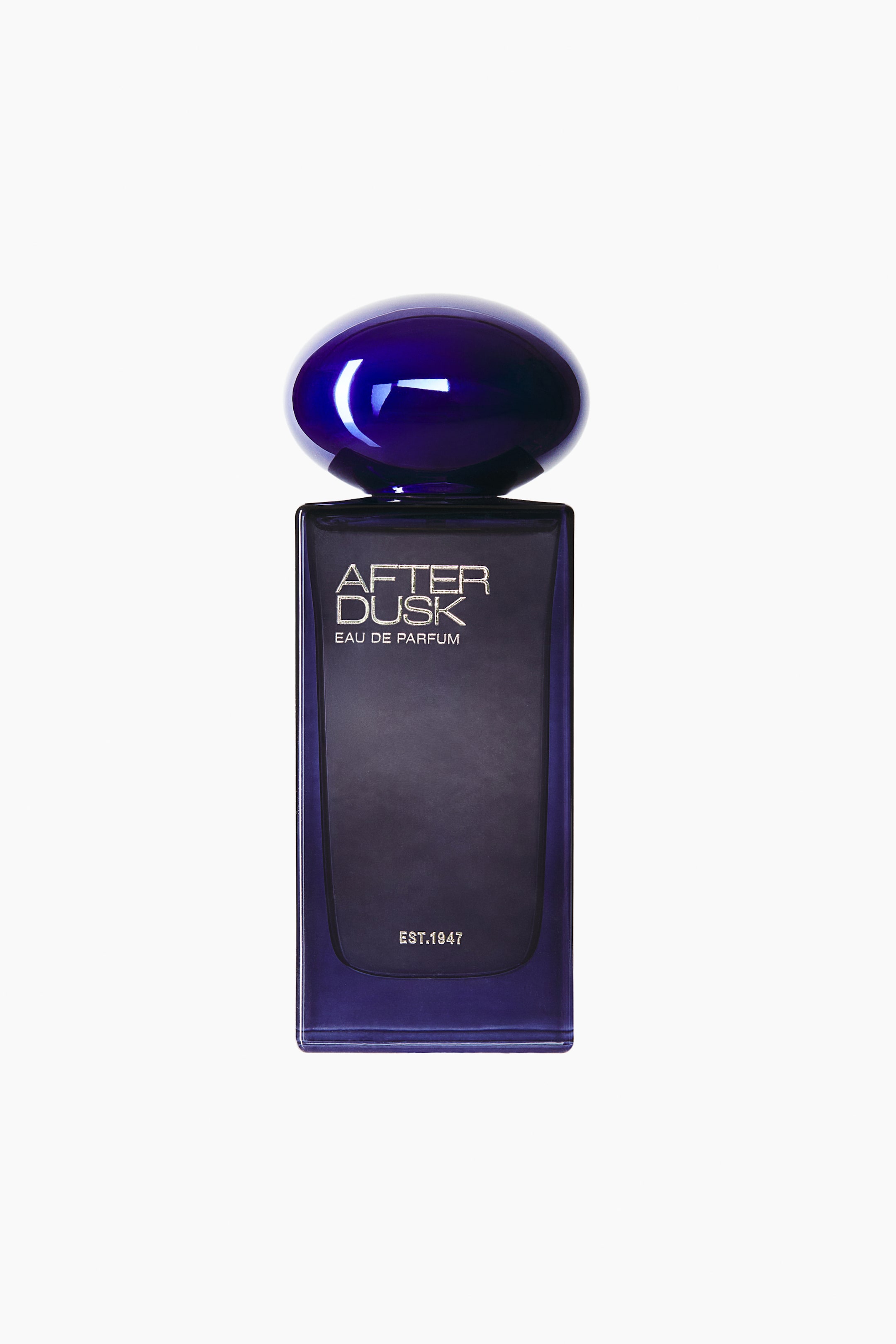 View larger image: A rectangular, dark purple glass perfume bottle stands upright in a front view, featuring a glossy, rounded oval cap in a matching deep purple hue. The front of the translucent bottle displays silver text reading "AFTER DUSK EAU DE PARFUM" near the top and "EST.1947" near the bottom.
