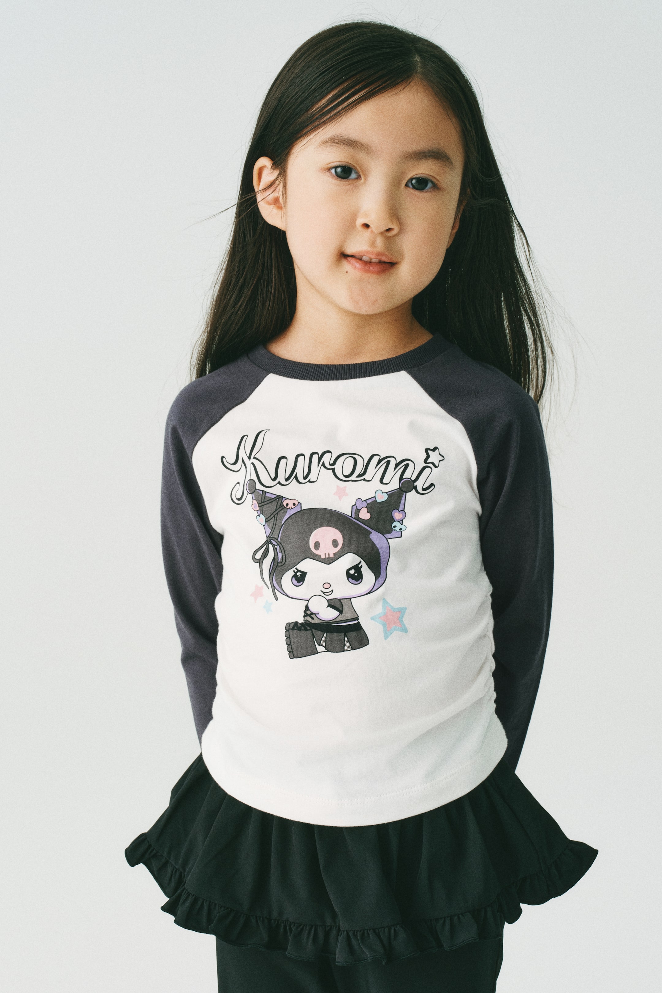 拡大画像を表示: モデルが着用しているのは、柔らかなプリント入りコットンジャージートップスです。丸いリブトリム付きのネックライン、ダークグレーのラグランスリーブ、そしてキャラクタープリントが施された白いボディが特徴です。サイドシームがギャザーになっており、トップスにシャーリング効果を生み出しています。このトップスは、黒のフリルスカートとスタイリングされています。