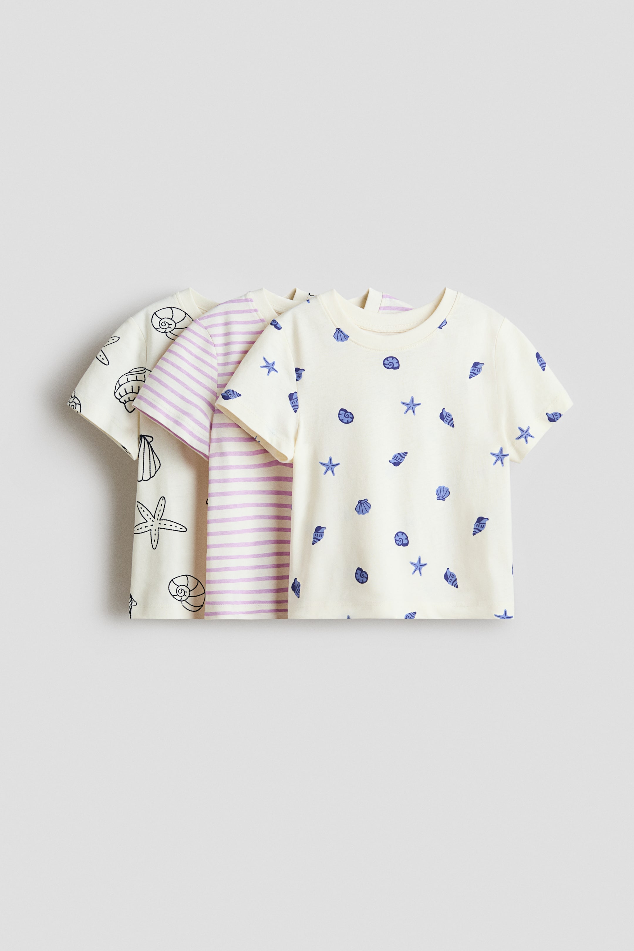 拡大画像を表示: 3枚のプリントされたコットンジャージーTシャツがそれぞれの前面を見せています。各Tシャツはリブネックラインとやや落ちたショルダーを特徴としています。最前面のTシャツは、クリーム色のベースに青い貝殻とヒトデのデザインを見せており、中央のTシャツは淡い紫と白のストライプを特徴とし、背面のTシャツはクリーム色のベースに黒のアウトラインで描かれた貝殻とヒトデを展示しています。