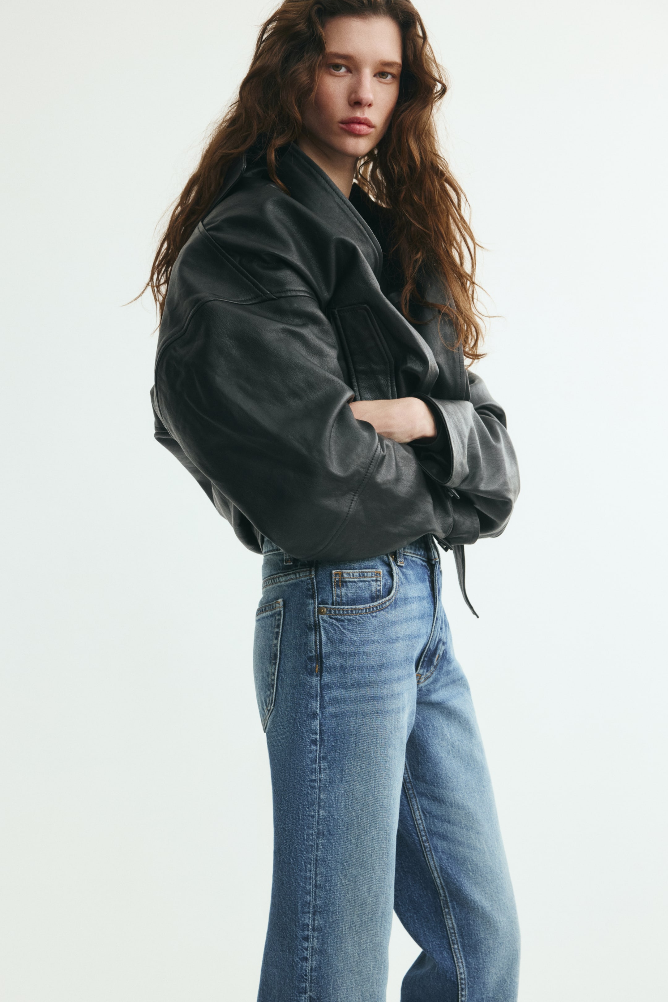 Xem ảnh lớn hơn: Người mẫu đang mặc quần jeans denim cạp cao màu xanh, phối cùng áo khoác da đen. Quần jeans có thiết kế cổ điển 5 túi với đường chỉ may lộ rõ và phần cạp ôm vừa vặn.