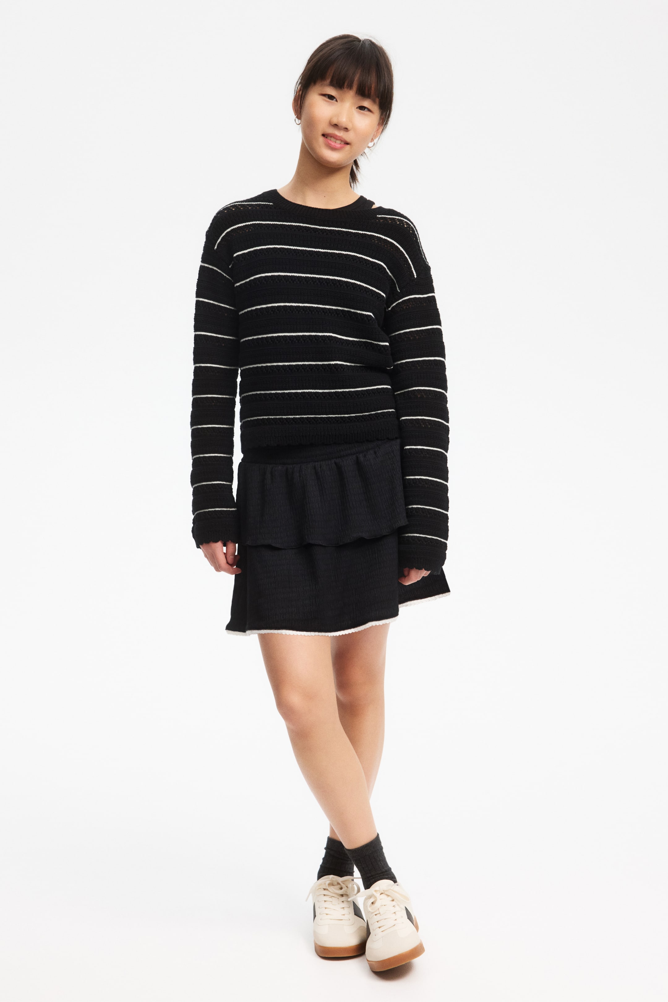 View larger image: A black crewneck sweater with horizontal white stripes and an open-knit construction is worn over a black tiered mini skirt. The skirt features a ruffled top tier and a lower tier finished with a white scalloped edge.