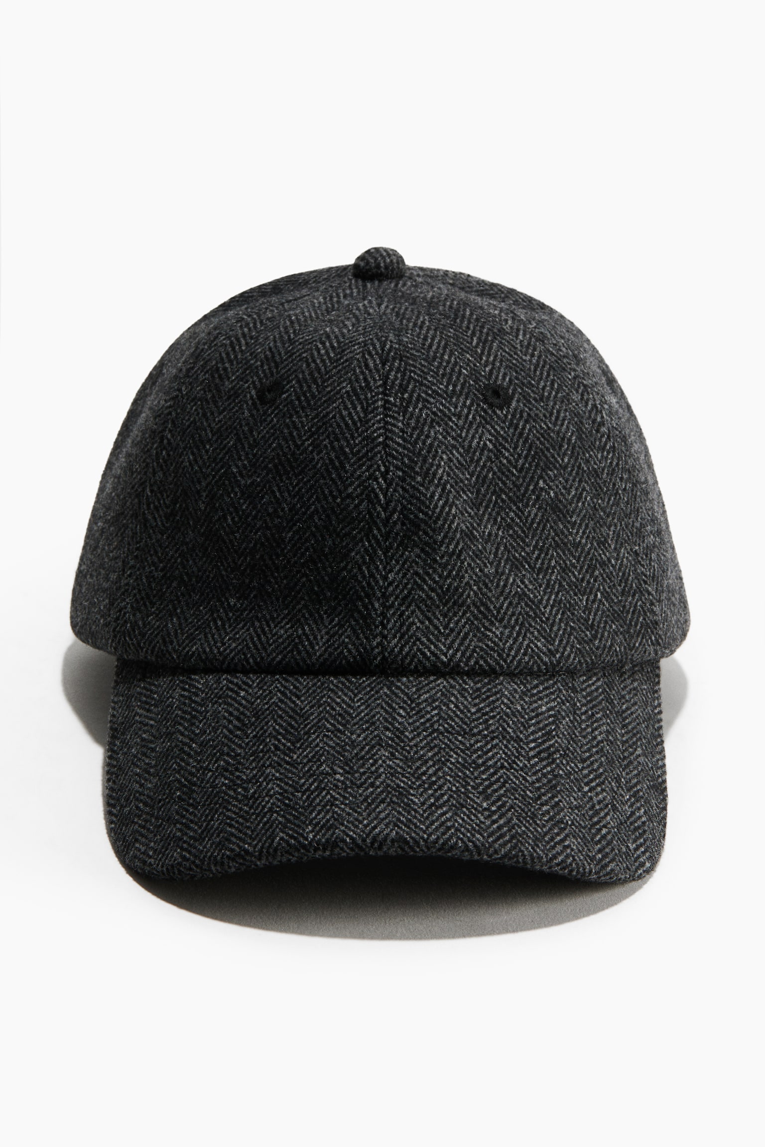 A dark grey cap in a soft herringbone-patterned fabric is shown from the front, displaying its curved peak and classic six-panel construction. The cap features a matching fabric-covered button at the top and embroidered ventilation eyelets.