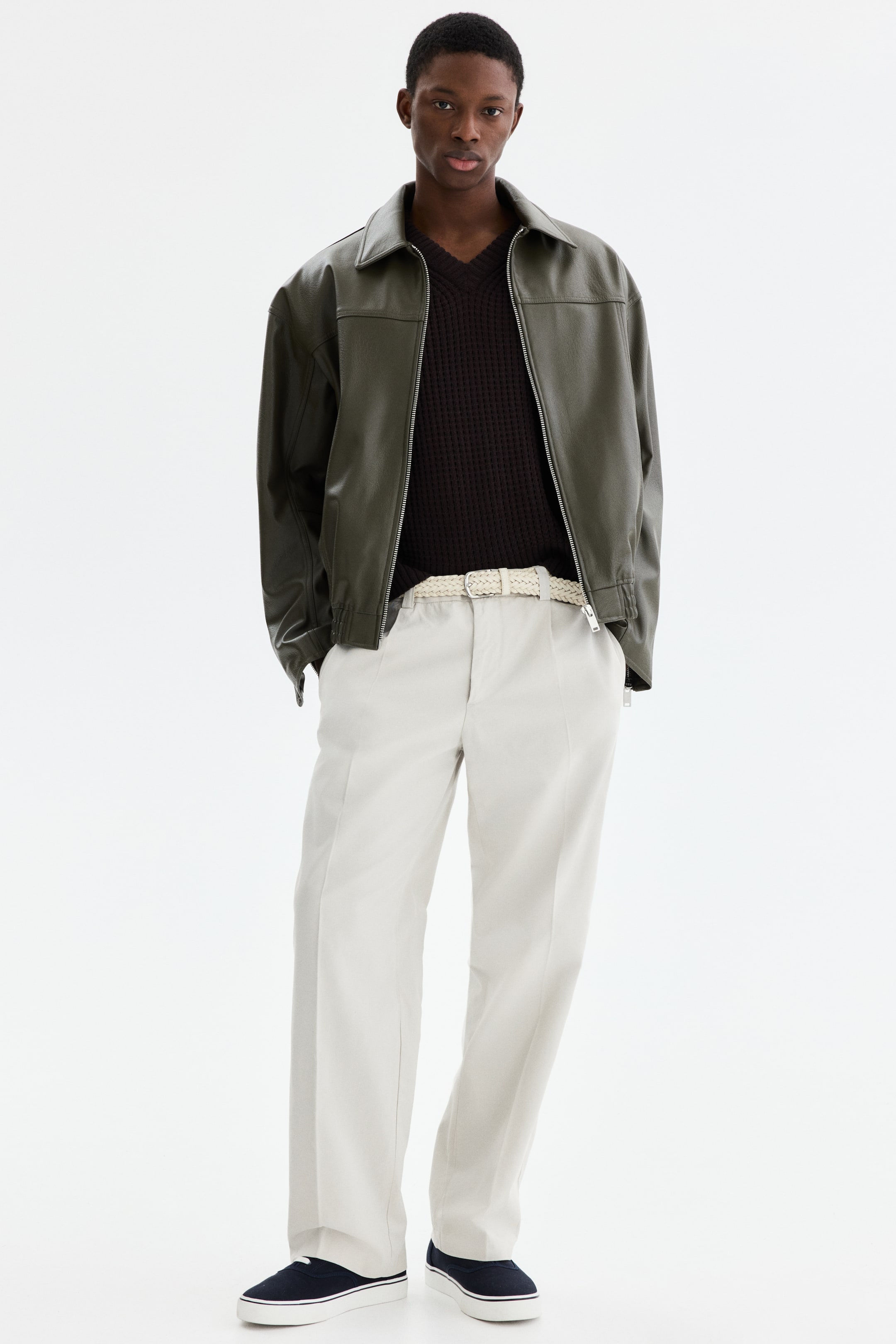 View larger image: The ensemble showcases a dark olive green bomber jacket, styled open over a dark brown V-neck knit jumper. The look is rounded off with light beige straight-leg trousers, secured by a cream braided belt, and navy slip-on runners.