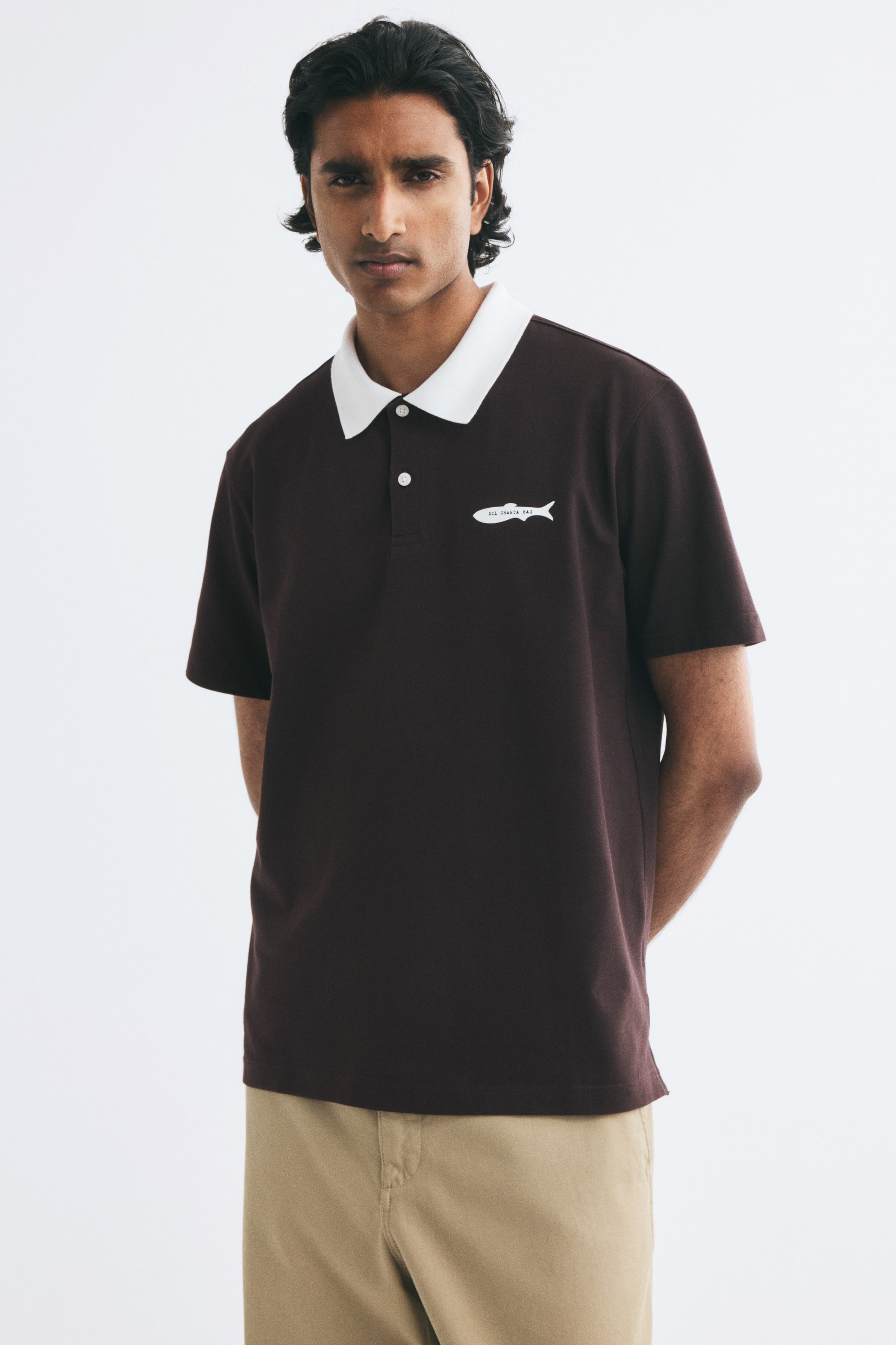 View larger image: The model is wearing a dark brown polo shirt with a contrasting white collar and a small white fish graphic on the left chest. The short-sleeved shirt features a two-button placket and is styled with beige trousers.