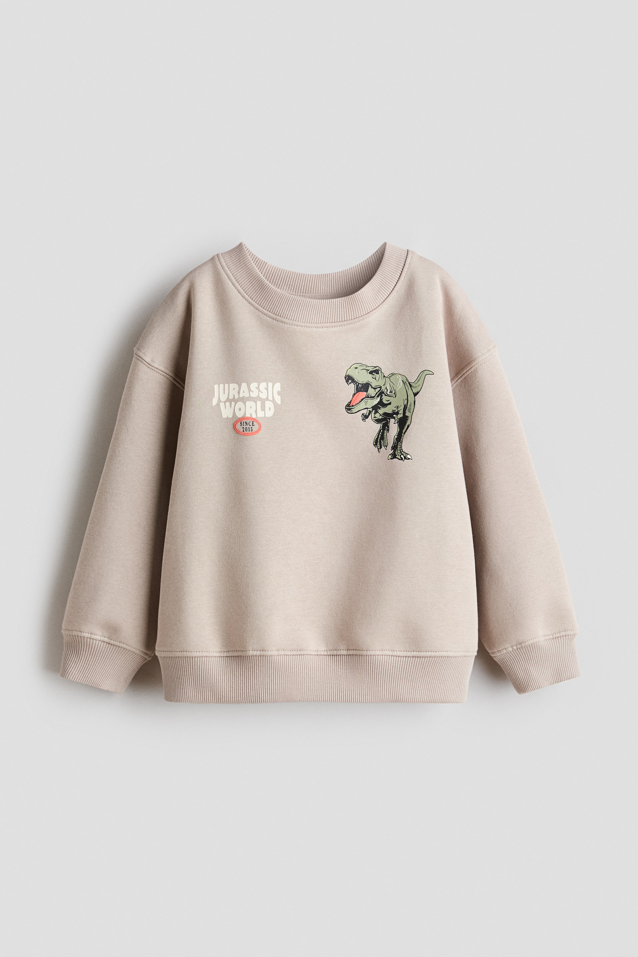 Zobrazit větší obrázek: Světle béžový top z potištěného mikinového materiálu je zobrazen zepředu; na přední straně nese textovou grafiku 'JURASSIC WORLD' s nápisem 'SINCE 2015' pod ní a zelenou grafiku dinosaura Tyrannosaurus Rex na hrudi. Je navržen s kulatým výstřihem s žebrovaným lemem, sníženými rameny, dlouhými rukávy a žebrovanými manžetami a spodním lemem.