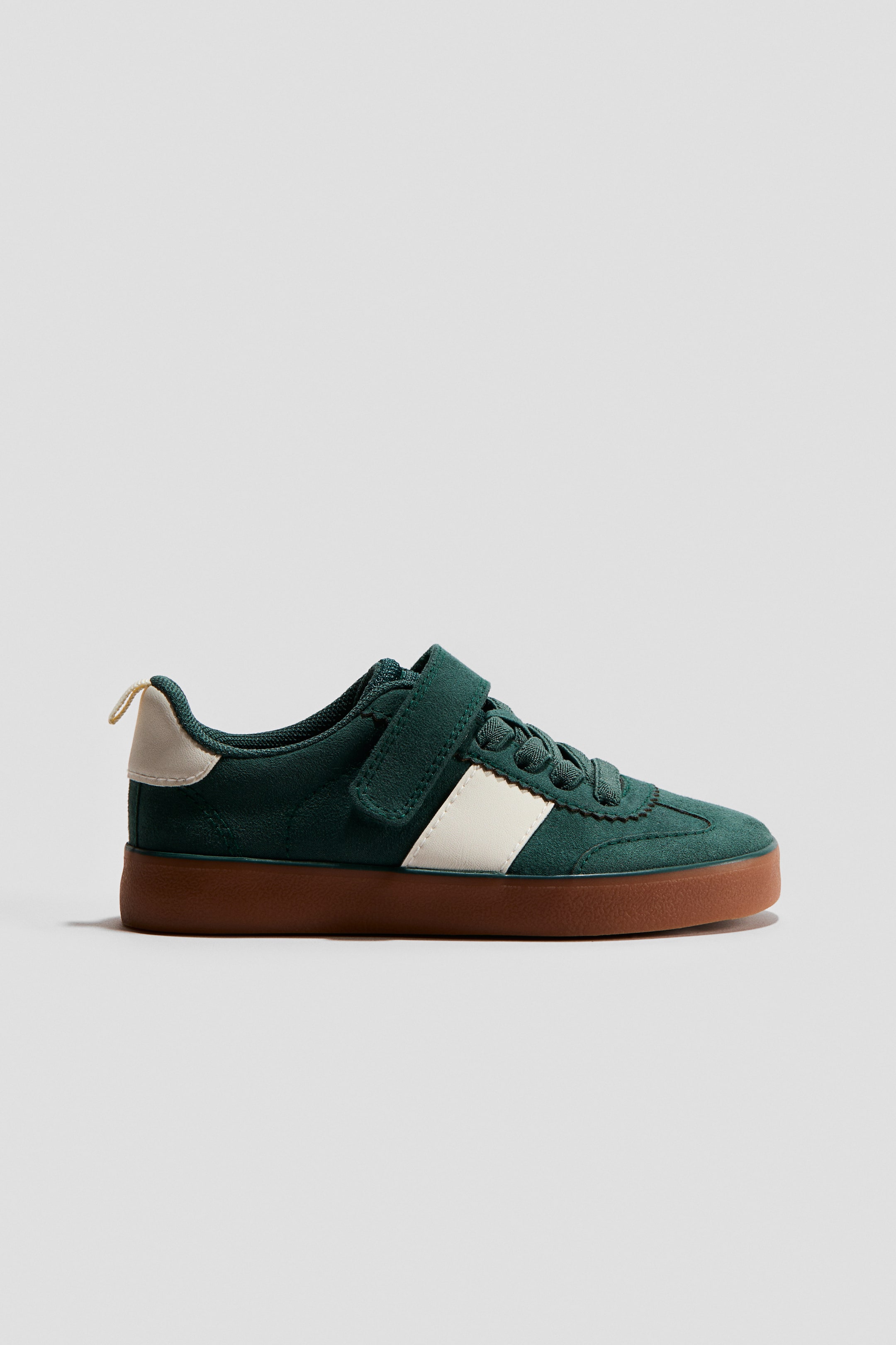 Zobraziť väčší obrázok: Tmavozelená teniska má vrchnú časť pripomínajúcu semiš s bočným pásikom kremovej farby, jedným pásikom na suchý zips cez farebne zladené šnúrky a svetlý béžový dôraz na päte. Hrubá hnedá podrážka tvorí základ topánky.