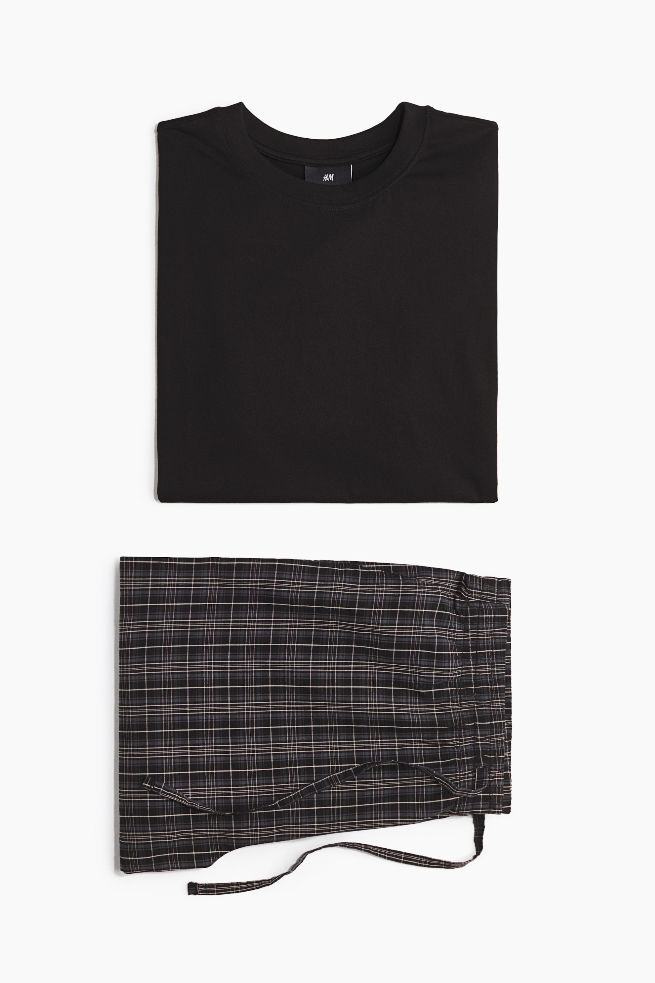 View larger image: Pyjamas show a folded black T-shirt with a round, rib-trimmed neckline, placed above folded dark plaid pyjama bottoms with a visible drawstring.
