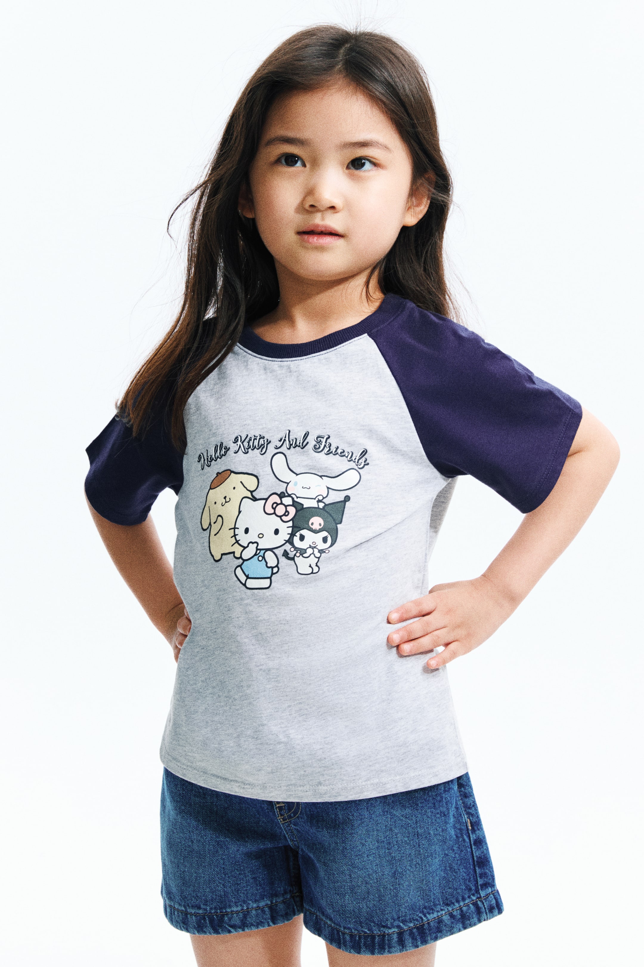 拡大画像を表示: Tシャツは、ライトグレーのボディにダークブルーのラグランショートスリーブとクルーネックラインを備えています。フロントには「ハローキティとフレンズ」のグラフィックプリントが施され、ポムポムプリン、ハローキティ、シナモロール、クロミが描かれています。ダークブルーのデニムショーツにゆったりとフィットします。
