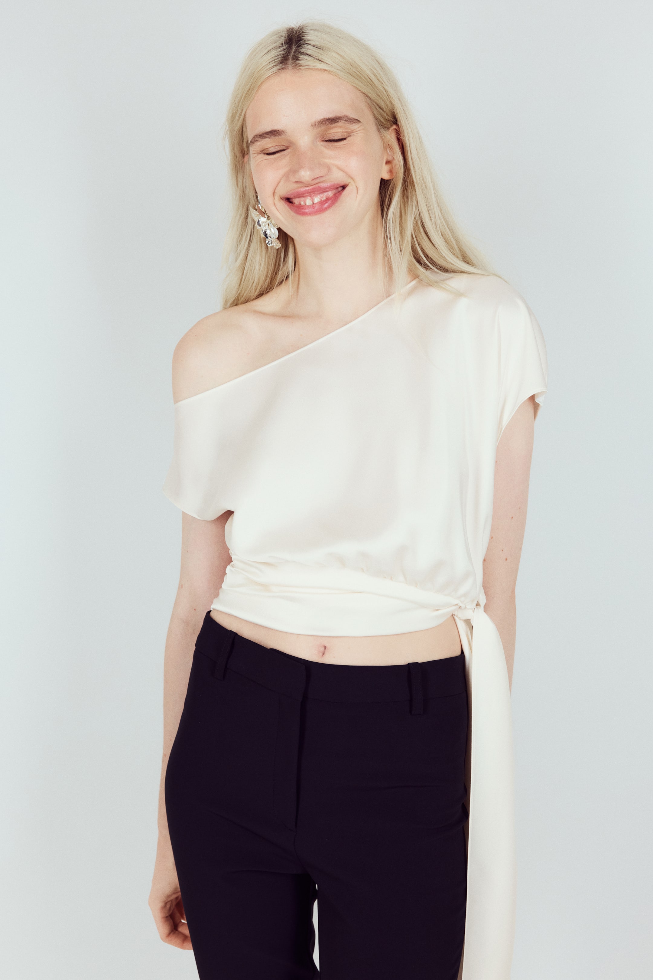 View larger image: A cream-coloured, shiny, cropped, one-shoulder wrap top is being worn, angled slightly to the right. It features a short sleeve on the right side and a tie closure at the left waist, forming a long sash extending downwards.