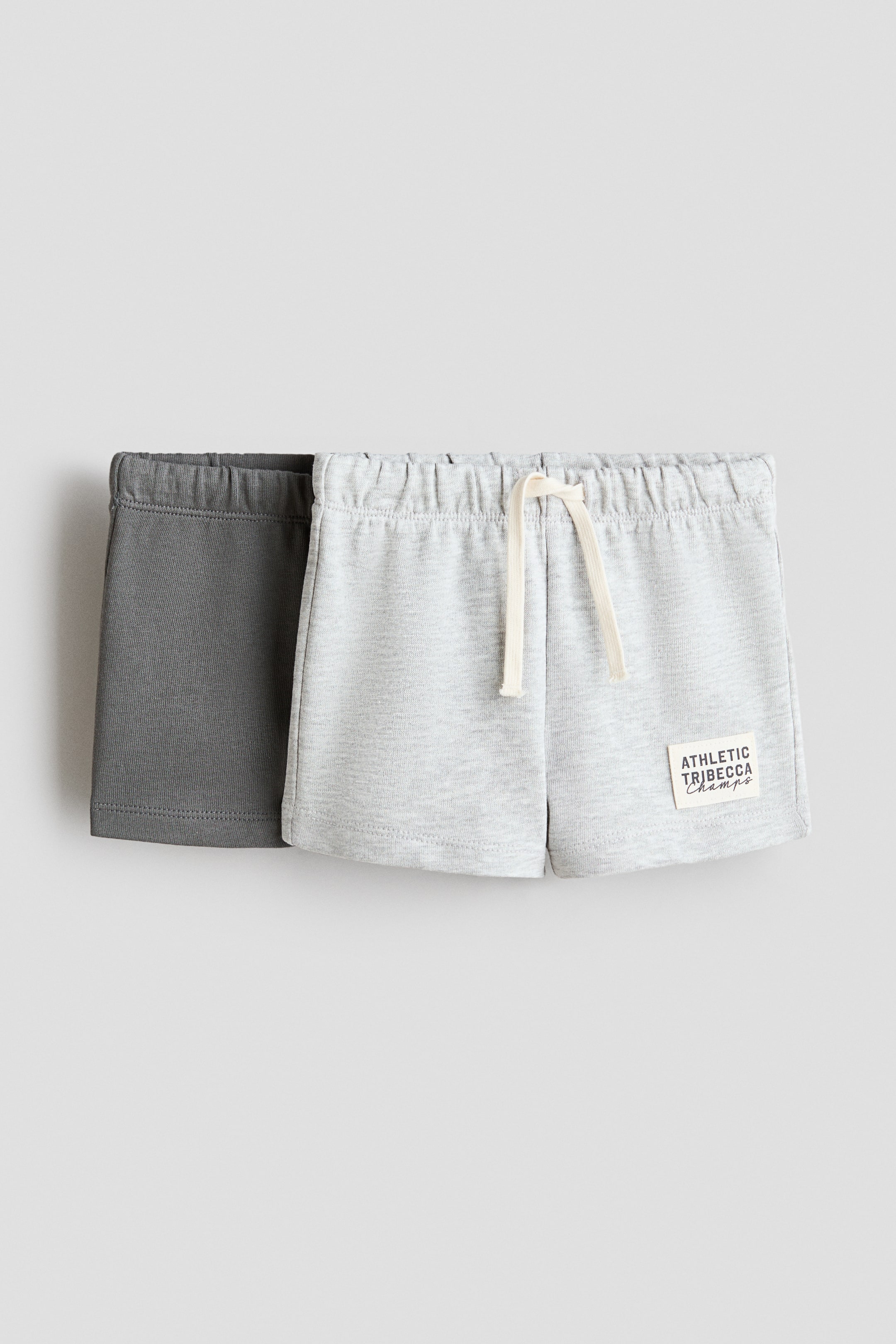 View larger image: Two pairs of casual shorts are showcased from the FRONT, with a light grey pair partially layered over a dark grey pair. The front light grey short features an elastic waistband with an off-white drawstring and a rectangular patch on the lower left leg that reads "ATHLETIC TRIBECCA Champs".