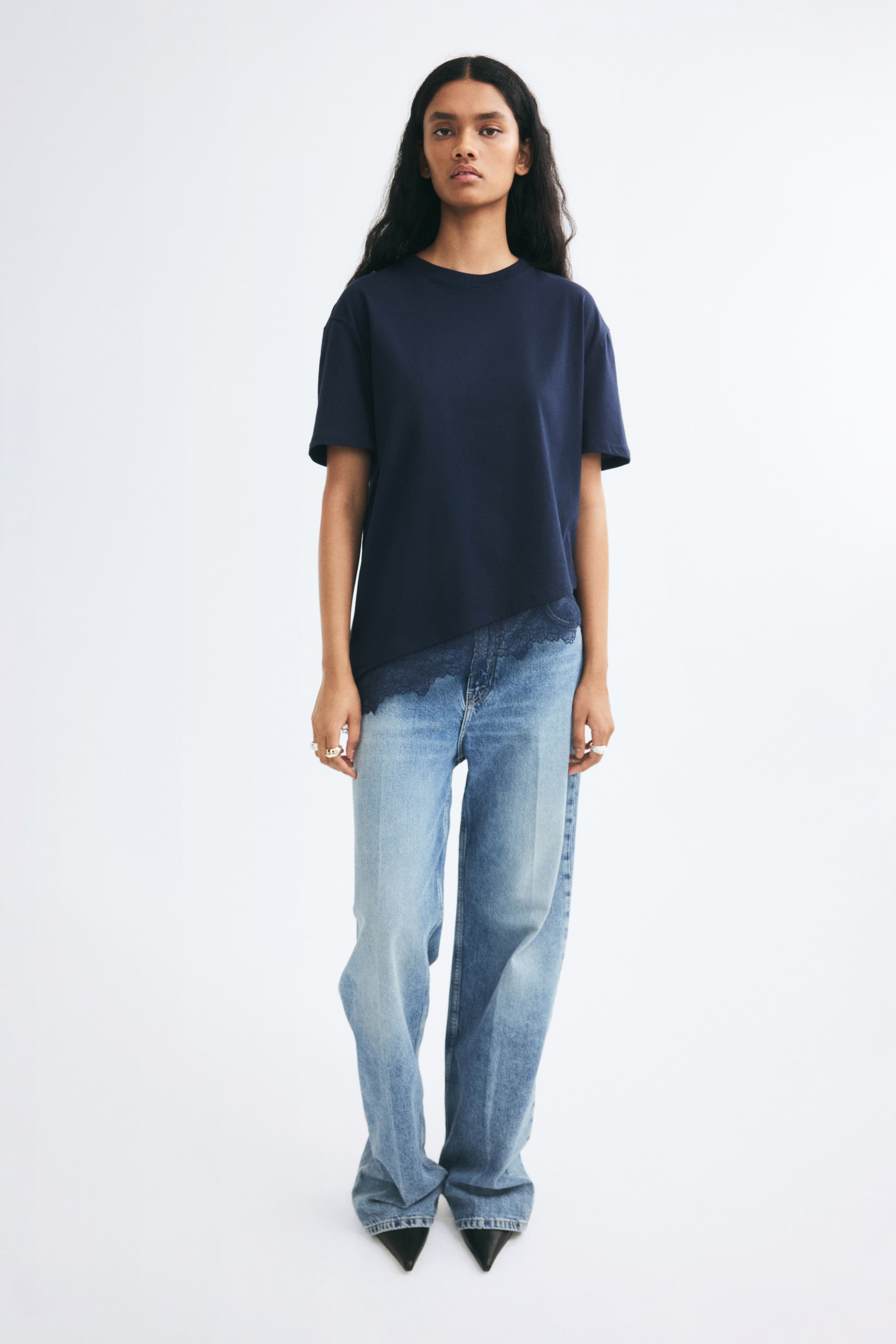 View larger image: A navy blue relaxed-fit T-shirt is being worn by the model, featuring dropped shoulders, a ribbed crew neckline, and an asymmetrical hem with scalloped lace trim. The T-shirt is styled loose over light-wash, wide-leg jeans.