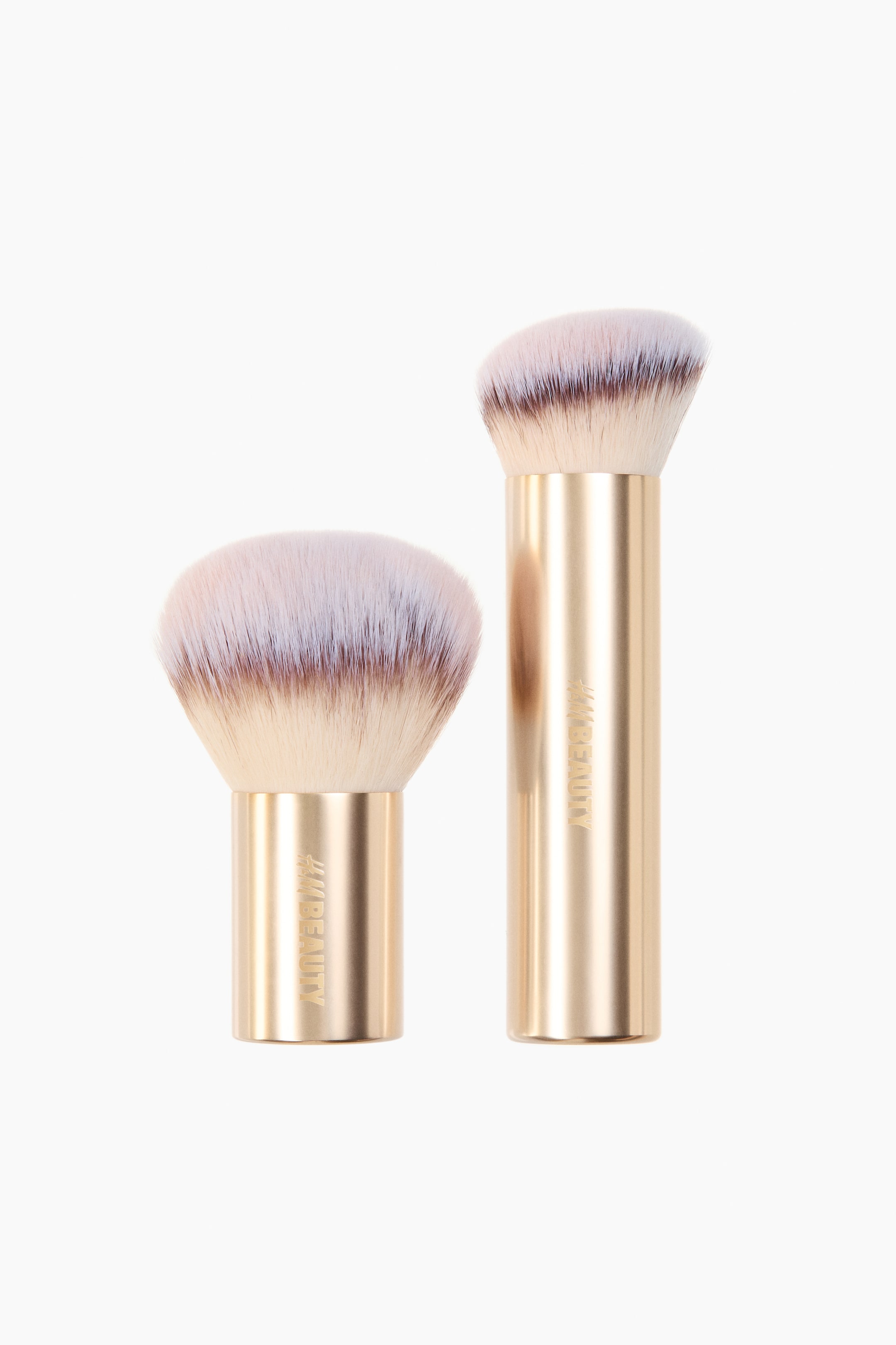 View larger image: A pair of bronzer brushes is shown, each featuring a golden cylindrical handle with '#HM BEAUTY' branding visible on the front. The taller brush on the right has an angled head with dense, two-tone bristles, while the shorter brush on the left has a rounded kabuki head with fluffy, two-tone bristles.