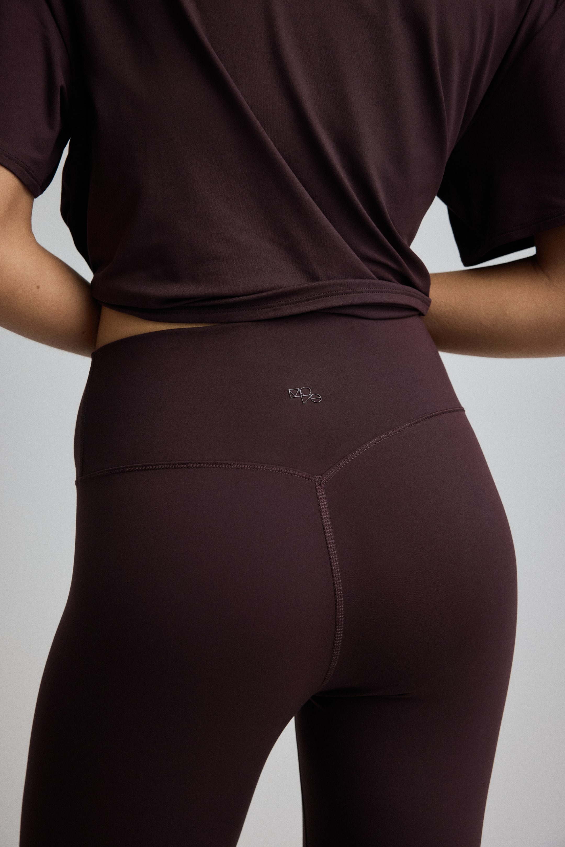 View larger image: Deep plum leggings are being worn from a back angle, featuring a high waist and a V-shaped seam detail on the back waistband. A small metallic logo is centered on the back waistband. A coordinating short-sleeved top is being worn, with its hem twisted just above the leggings' waistband.