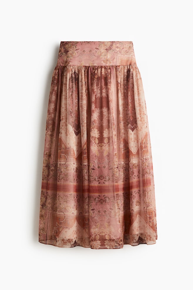 A front-facing long skirt is displaying a reddish-pink and brown ornate floral and paisley pattern. It is designed with a wide, flat waistband and a gathered, flowing silhouette in a sheer fabric. - 1