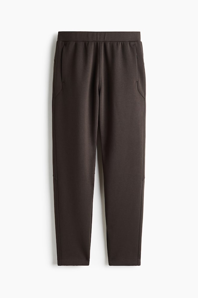 These dark brown knit pants are displaying a front view with a flat elastic waistband and angled side pockets. The legs are tapering toward clean-finished hems, highlighting a smooth, structured fabric. - 1