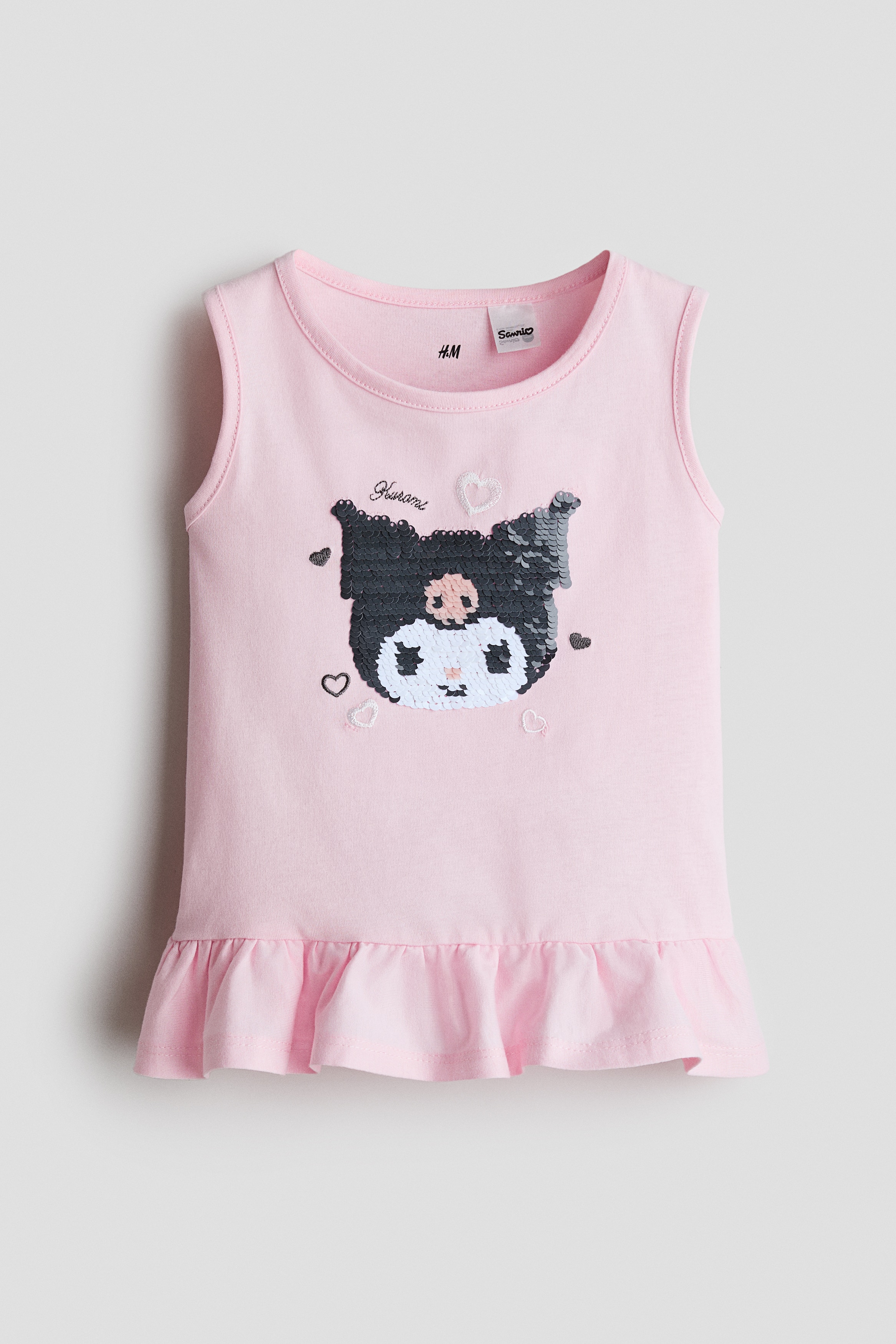 A light pink sleeveless top featuring a round neckline and a sequined character design on the front is worn by the model. The top has a flared frill trim at the hem and is styled with a matching pink tulle skirt.
