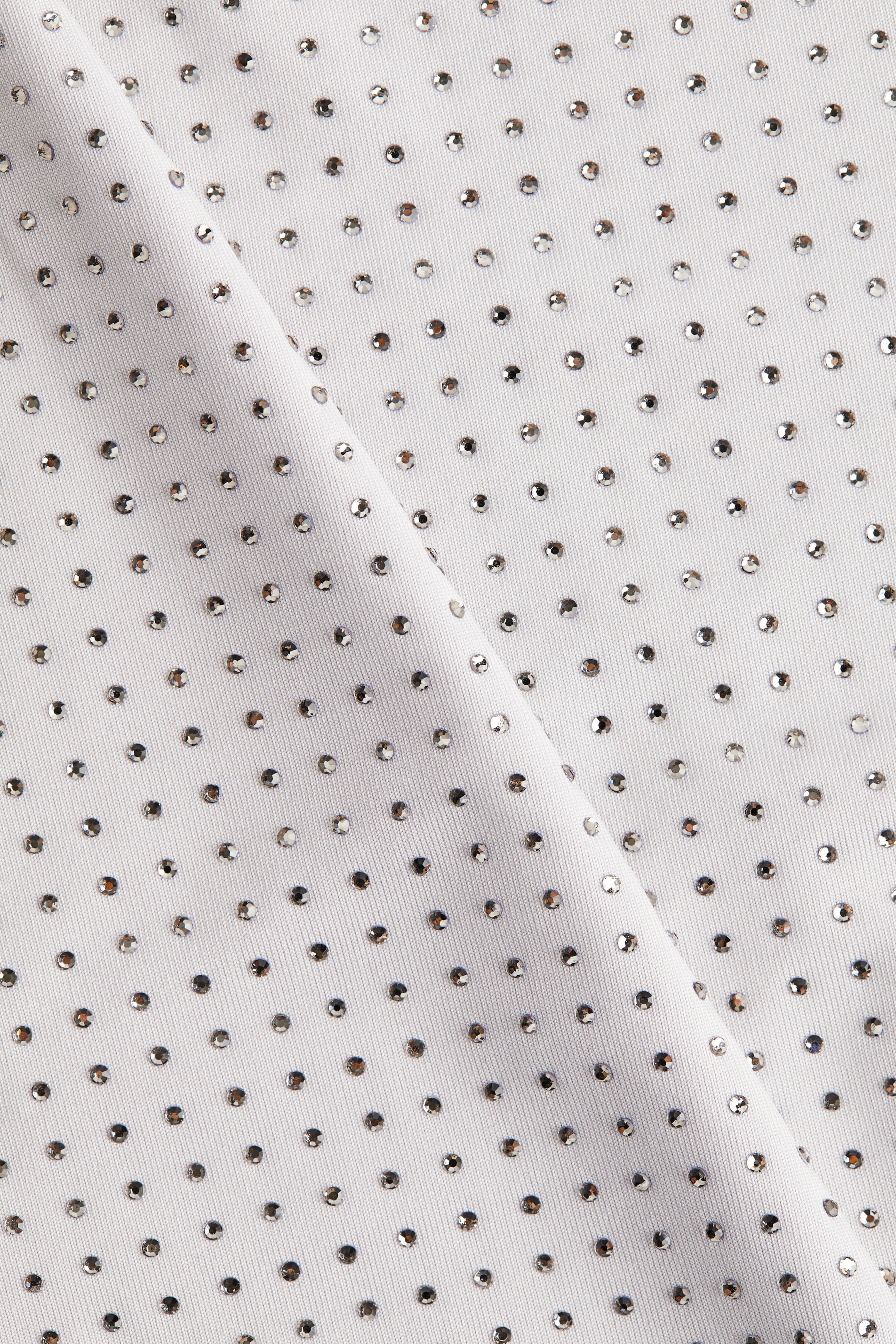 Grösseres Bild ansehen: Ein cremefarbener Stoff zeigt ein Muster aus gleichmäßig verteilten, kleinen, runden, silberfarbenen Strasssteinen. Der Stoff faltet sich sanft diagonal, wodurch subtile Schatten und Highlights auf seiner Oberfläche entstehen.