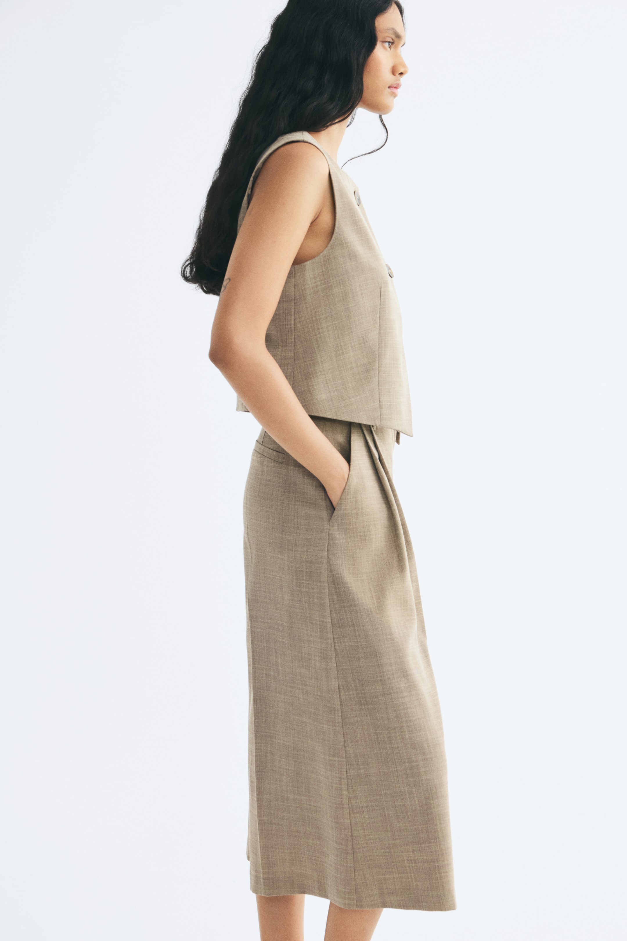View larger image: A beige, sleeveless woven vest featuring a round neckline and cropped hem is being worn by the model, who is standing in profile. The vest is paired with matching wide-leg trousers in the same fabric, with the model placing one hand in the trouser pocket.
