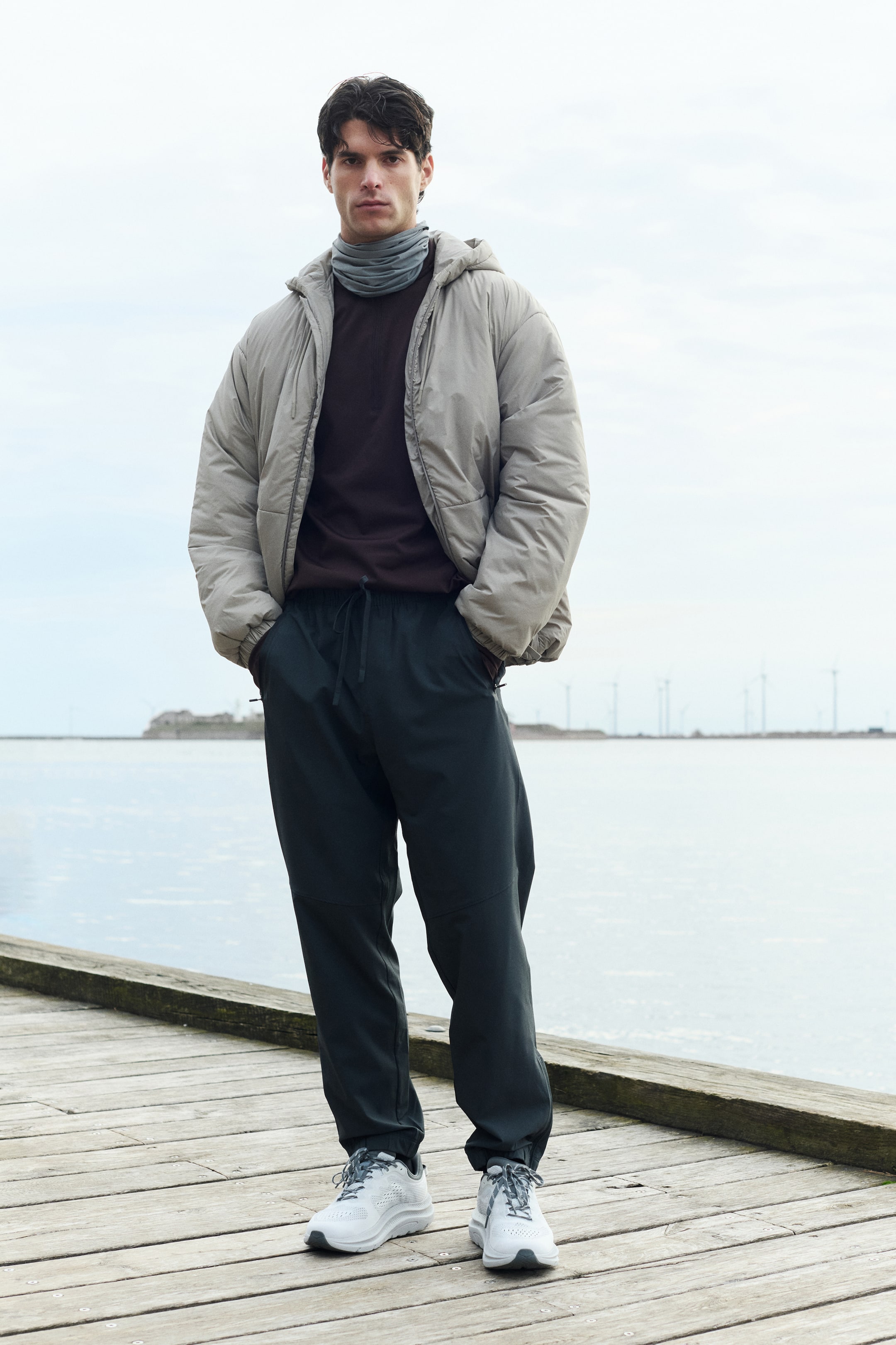Ampliar la imagen: Un conjunto para hombre presenta una chamarra puff con capucha en gris claro, una camisa de manga larga en marrón oscuro y un cuello de tortuga en gris. Unos pantalones de chándal en gris oscuro con un cordón visible y bolsillos con cierre complementan la parte superior, mientras que unos tenis deportivos en blanco y gris completan el look.