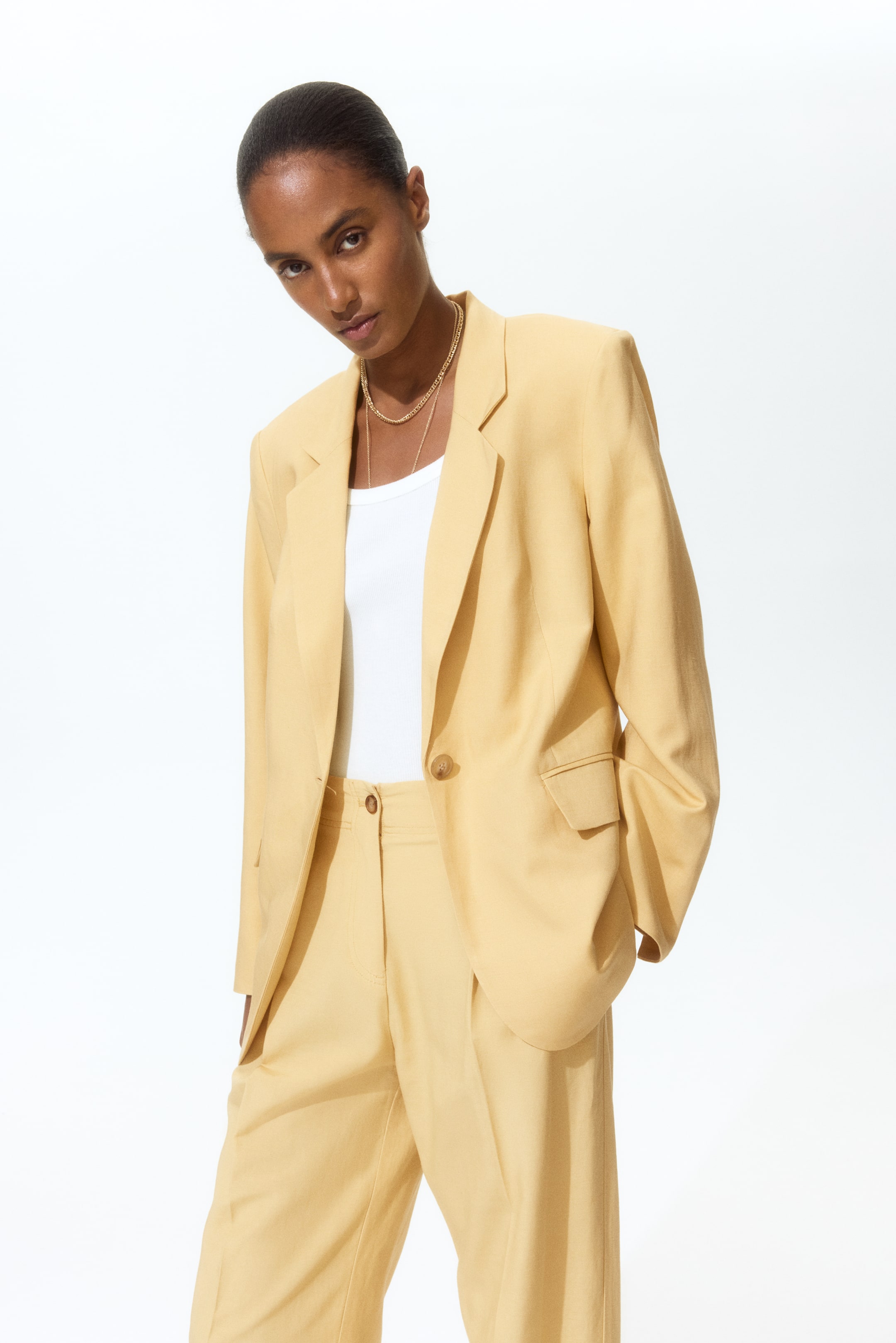 View larger image: A light yellow suit, featuring a single-breasted blazer with notch lapels, a one-button closure, and flap pockets, paired with matching high-waisted trousers, is showcased from a slightly angled front perspective. The blazer is elegantly layered over a white top, while the trousers are depicted with hands casually placed in their pockets.