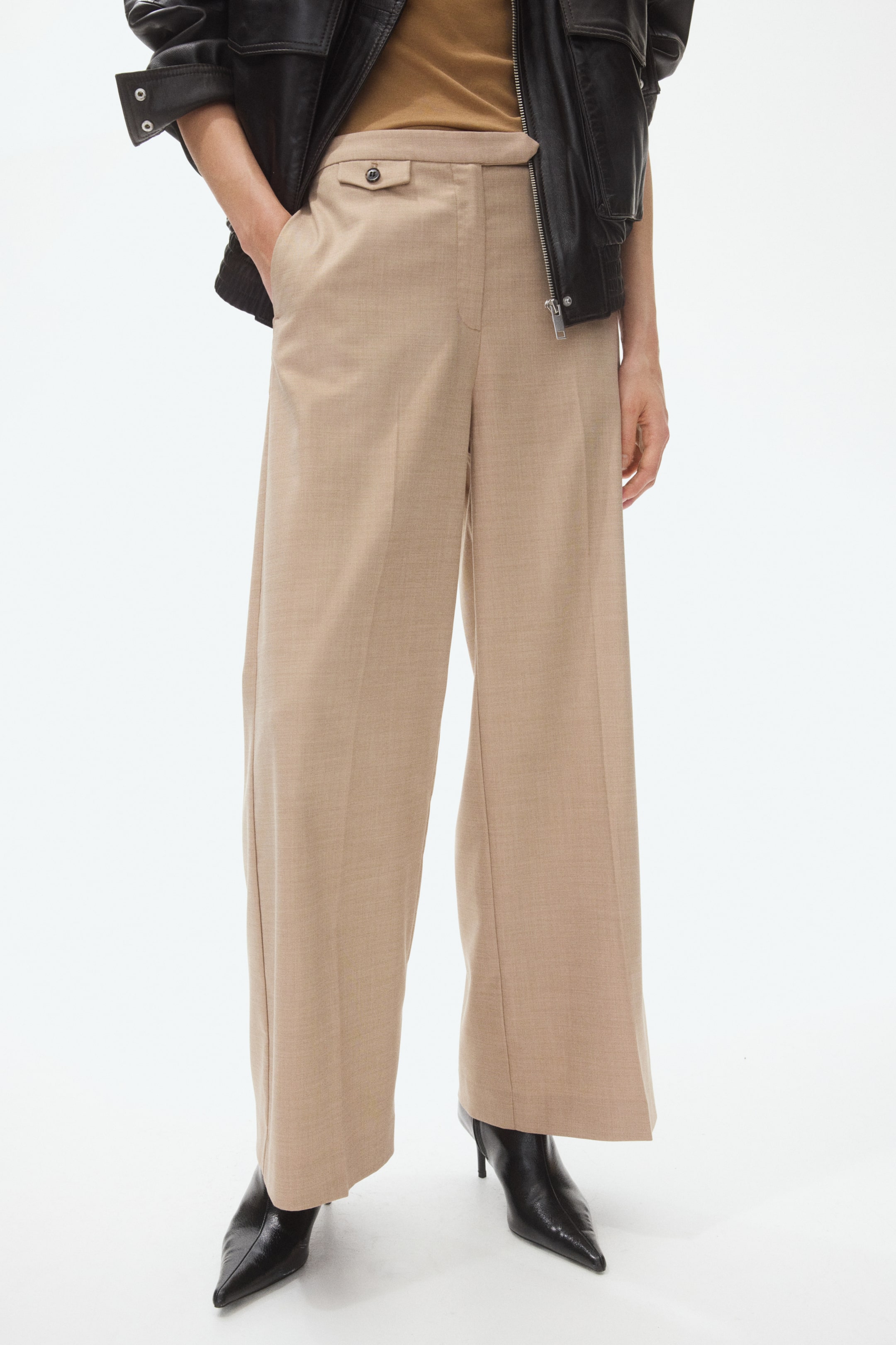 View larger image: Beige wide-leg trousers present a high-waisted fit, featuring a small buttoned flap pocket on the front right side. The trousers showcase a straight, wide leg with a prominent crease, and their hems fall over black pointed-toe boots.
