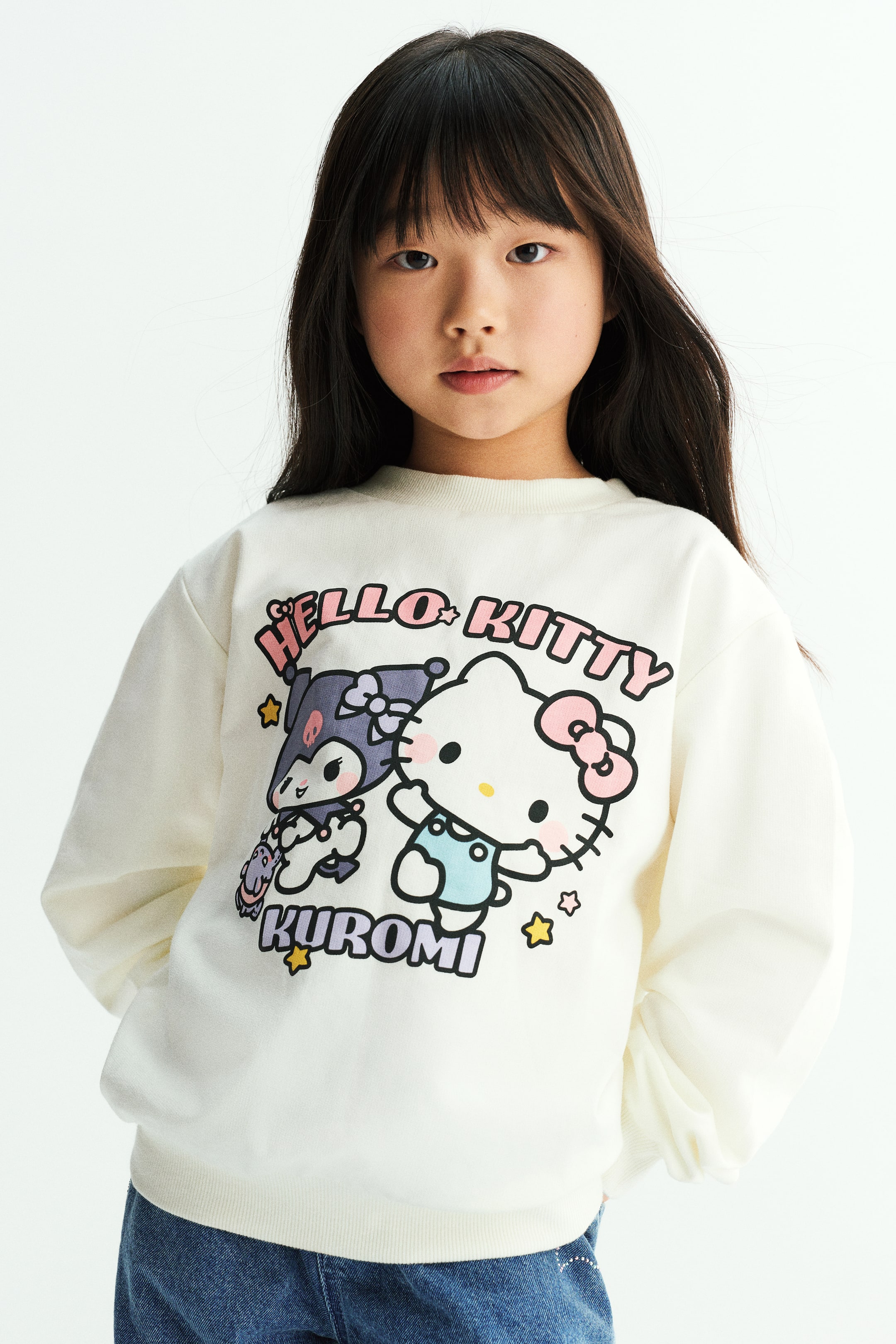 拡大画像を表示: クリーム色の長袖セーターをモデルが着用しており、フロントにはHello KittyとKuromiのカラフルなプリントグラフィックがあしらわれています。セーターは、リブ仕様のラウンドネック、ドロップショルダー、幅広のリブ裾が特徴で、ブルーデニムジーンズと合わせています。