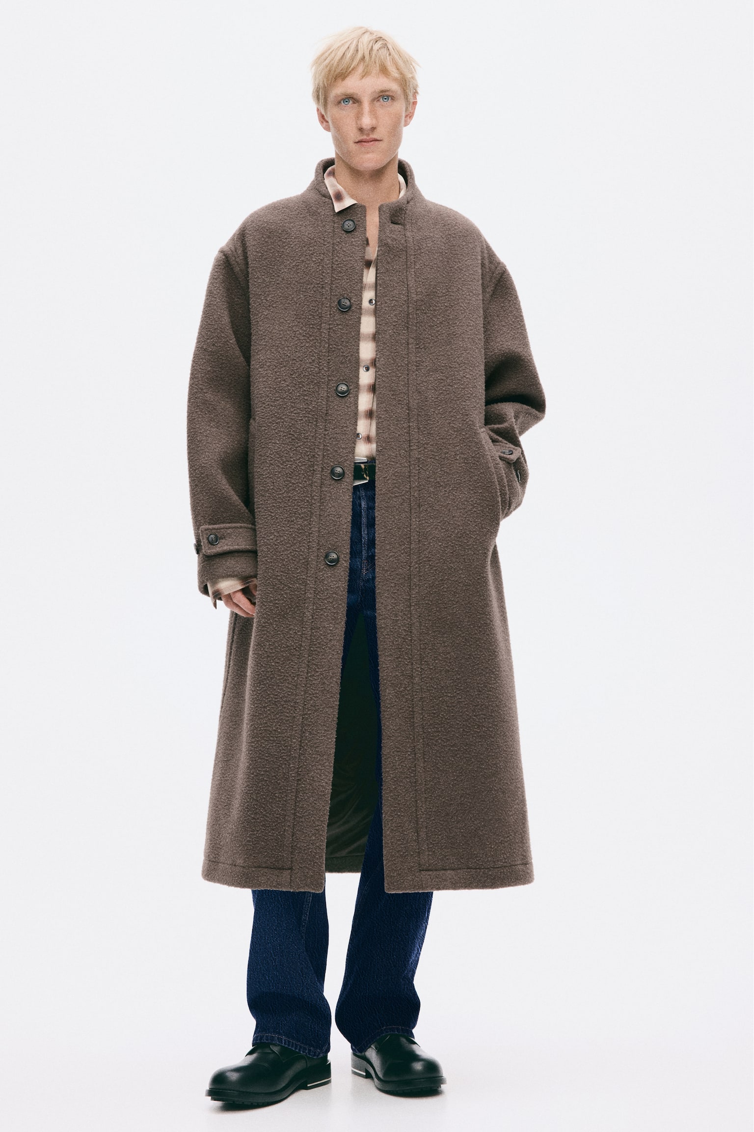 A long, textured brown wool-blend coat is being worn by the model, featuring a stand collar, front button fastening, and button-tab cuffs. The model is posing with one hand in a side pocket, layering the coat over a patterned collared shirt, dark blue jeans, and black shoes. - 1