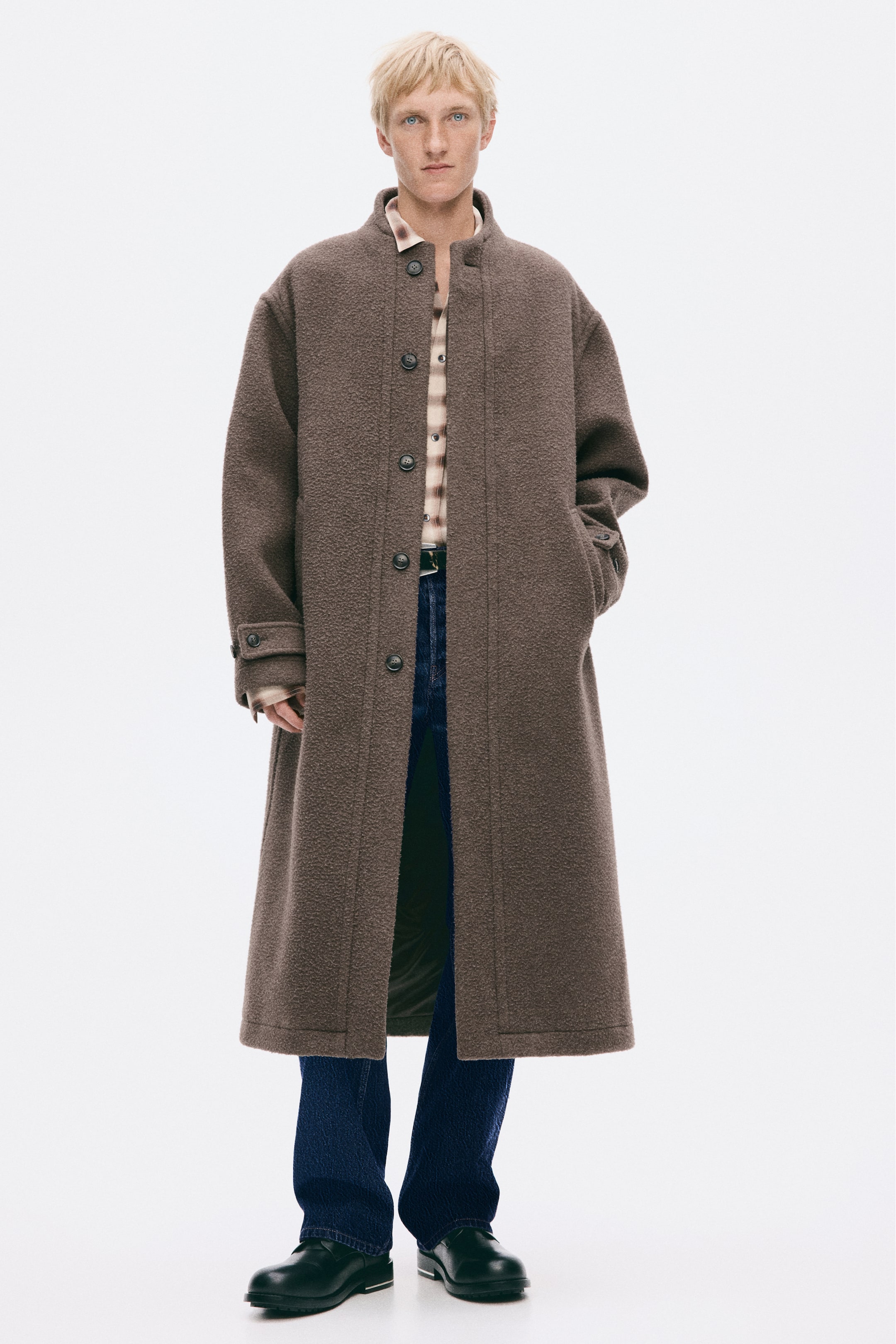 Visa större bild: En lång brun kappa i texturerad ullblandning bärs av modellen och har en ståkrage, knäppning framtill och ärmslut med knappflikar. Modellen poserar med ena handen i en sidoficka och bär kappan över en mönstrad skjorta med krage, mörkblå jeans och svarta skor.