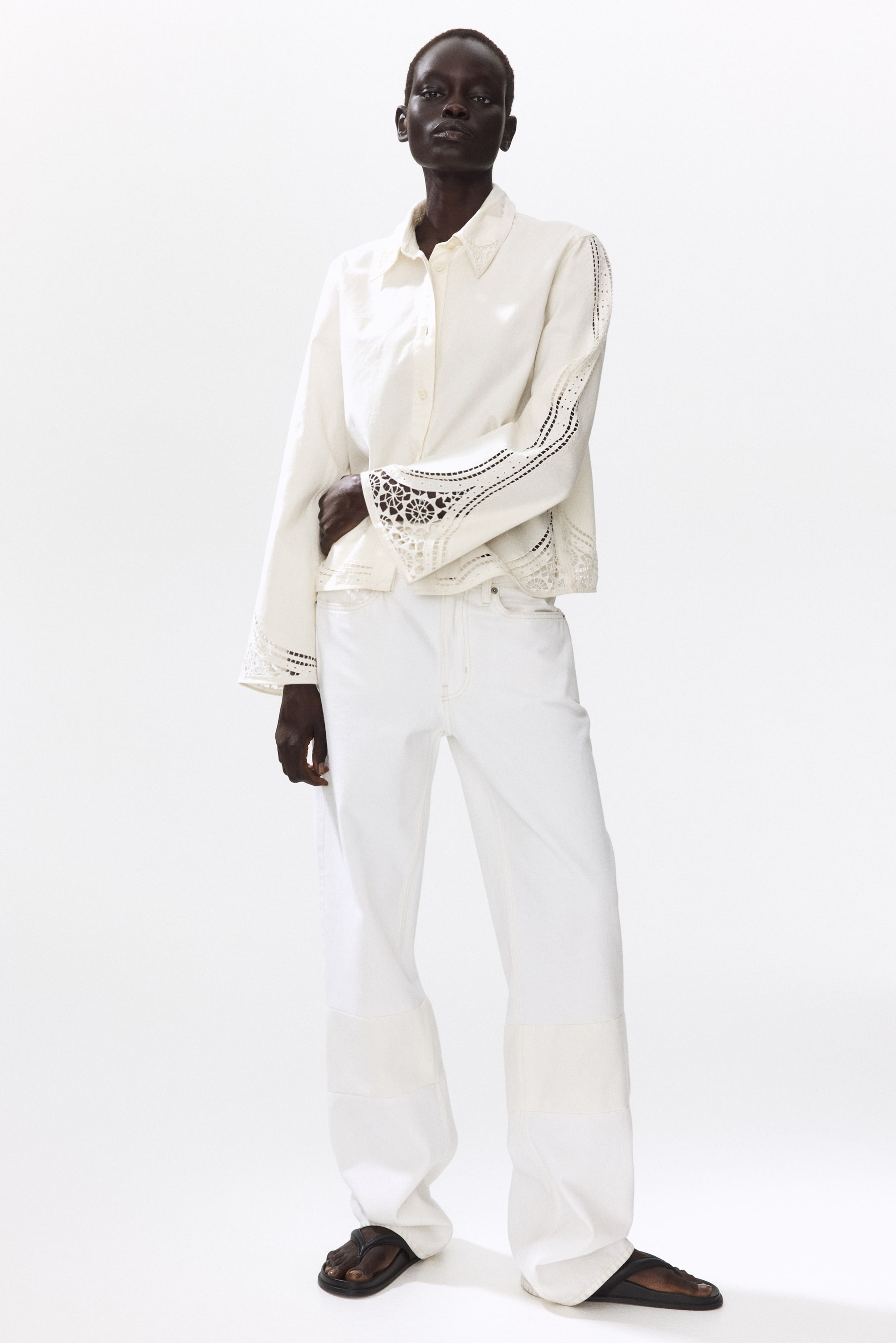 Agrandir l'image: Une veste courte en tissage de coton blanc cassé avec broderie anglaise est portée par le mannequin. Elle présente un col pointu et des boutons sur le devant. Le mannequin pose une main sur la veste, qui est stylée sur un pantalon blanc à jambes larges.
