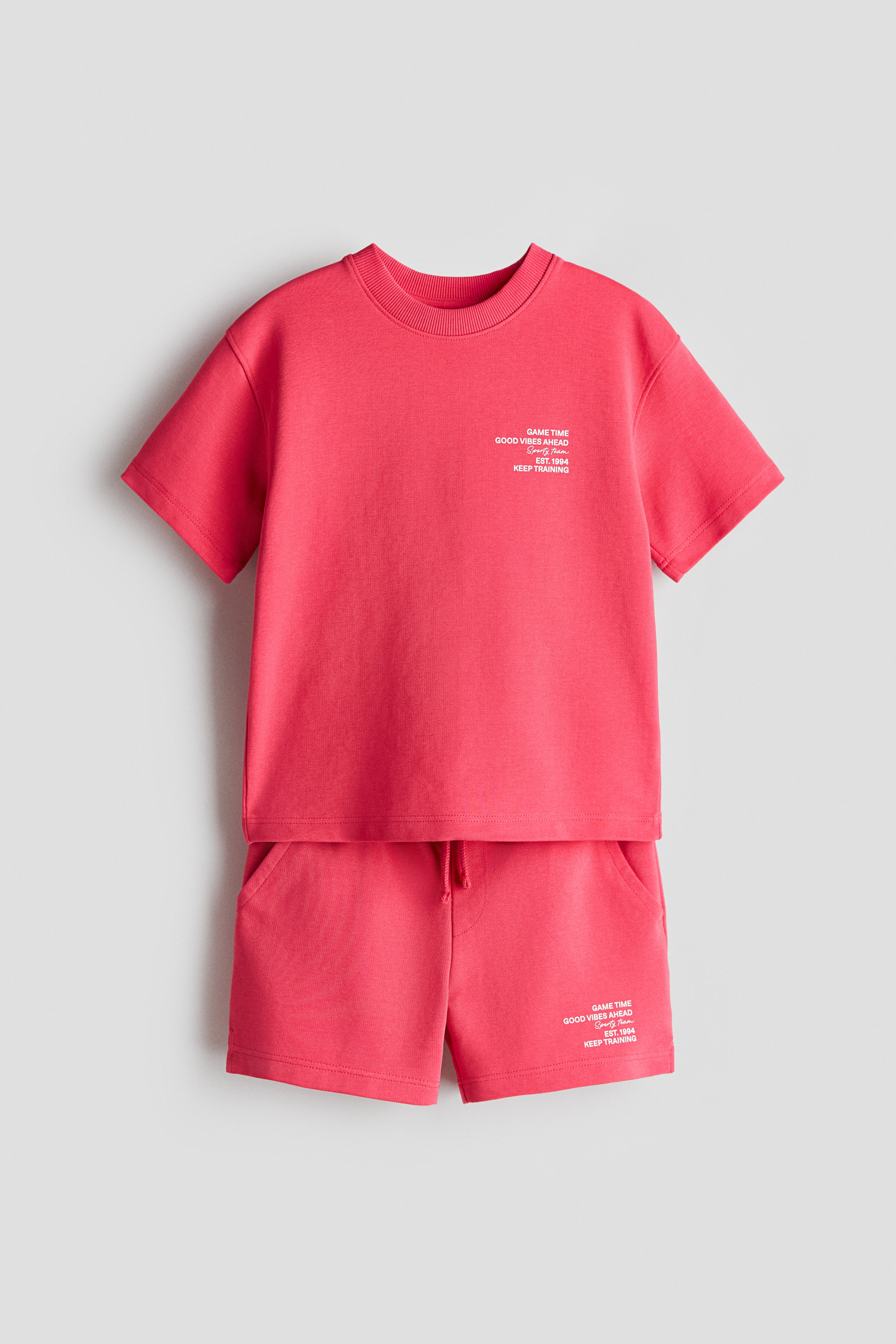 View larger image: A red two-piece set, comprising a short-sleeved top and matching shorts, is showcased FRONT. The top boasts a ribbed crew neck and short sleeves, adorned with white text graphics on the front. The shorts feature an elastic waistband with a drawstring, side pockets, and corresponding white text graphics on the front.