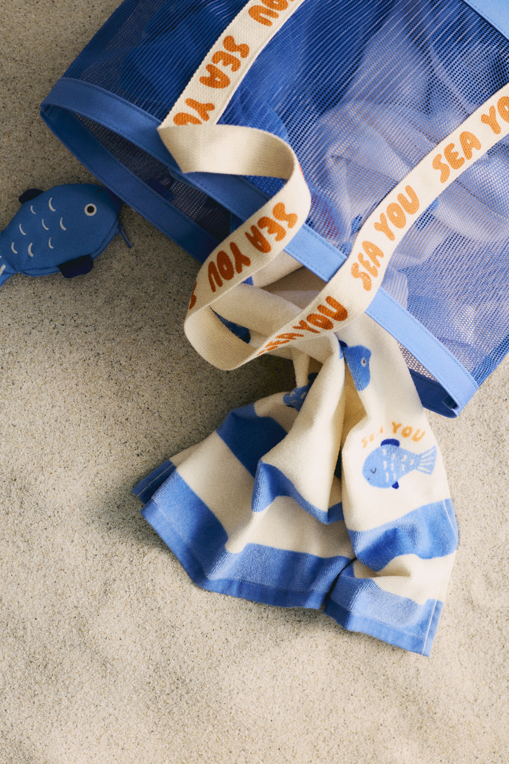 View larger image: A blue mesh tote bag, featuring a light blue fabric trim and cream straps with orange "SEA YOU" text, is angled from the top left. A blue and white wide-striped towel, displaying a blue fish graphic and "SEA YOU" text, is partially tucked into the bag, and a blue fish-shaped pouch with white scale details is lying next to it.