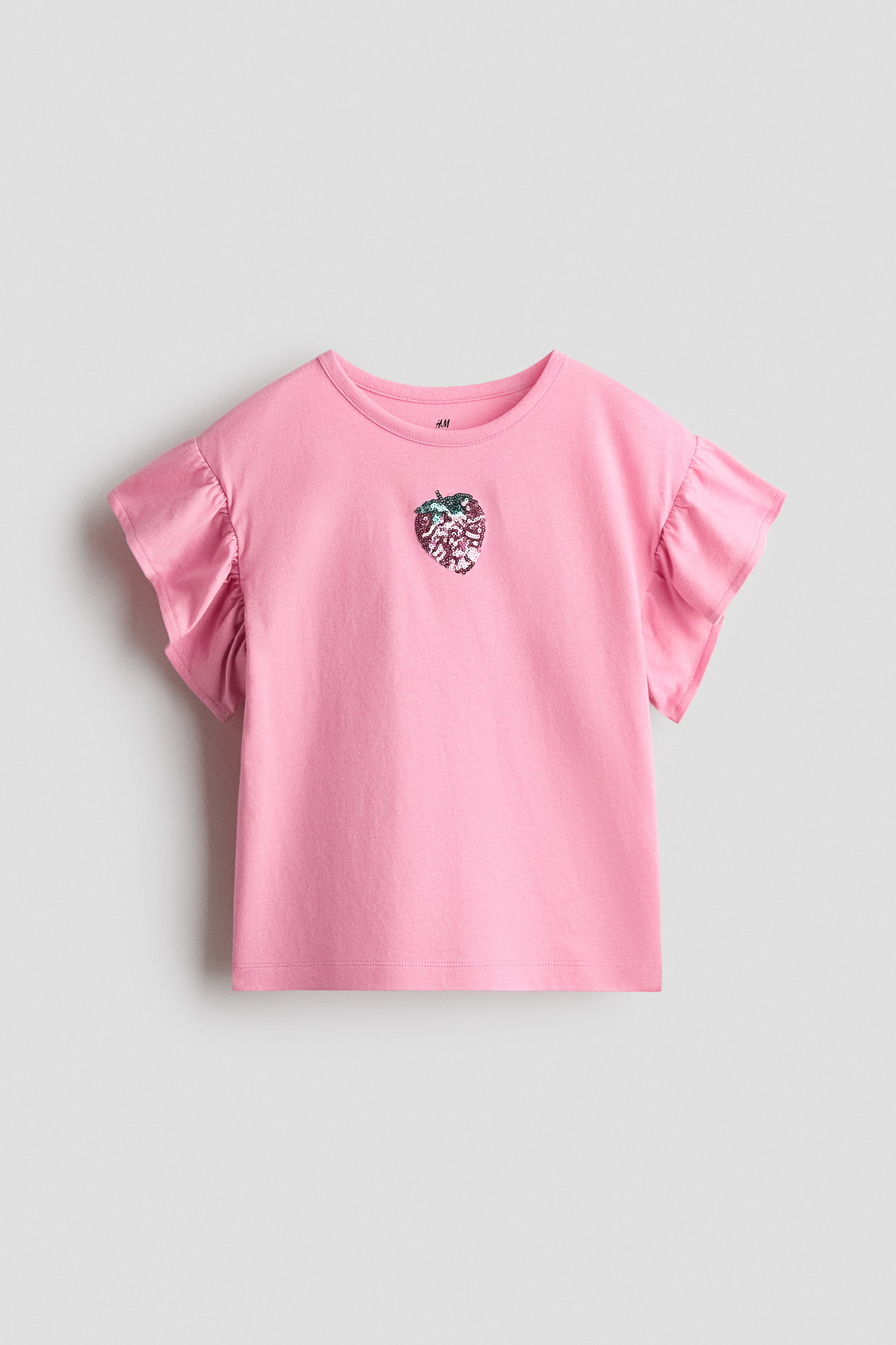 Näytä suurempi kuva: Vaaleanpunainen t-paita makaa tasaisena, siinä on pyöreä pääntie ja lyhyet, röyhelöiset hihat. Keskellä edessä on kimalteleva, monivärinen paljettimansikkakuvio.