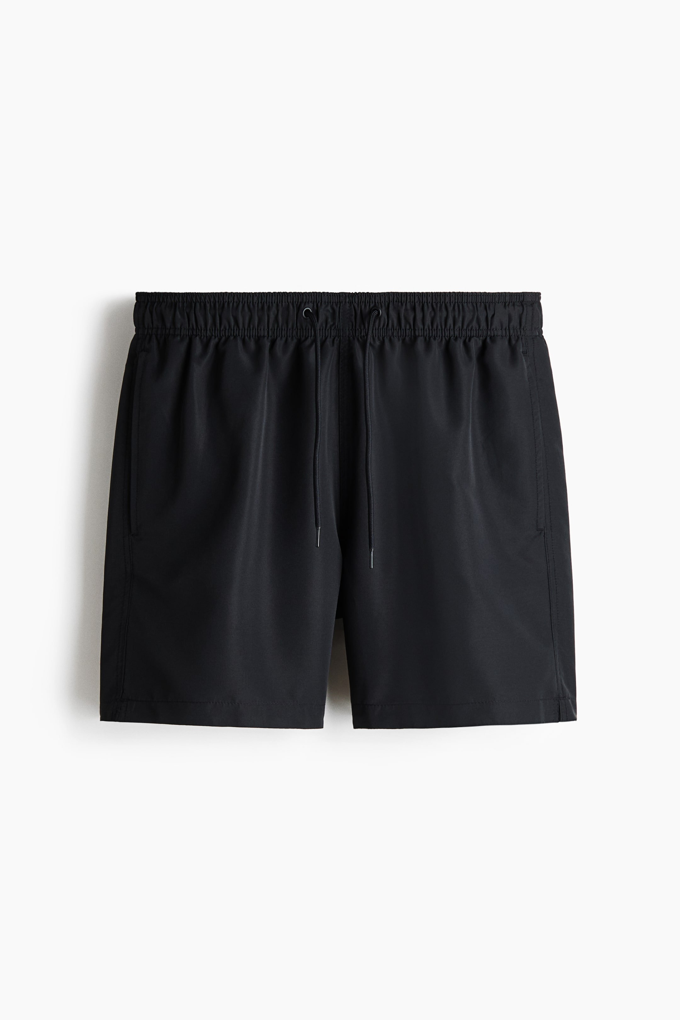 View larger image: Black shorts are lying flat, facing forward, featuring an elasticated drawstring waistband with black cords and silver aglets. Side seam pockets are visible on both sides, and small side vents are appearing at the outer leg hems.