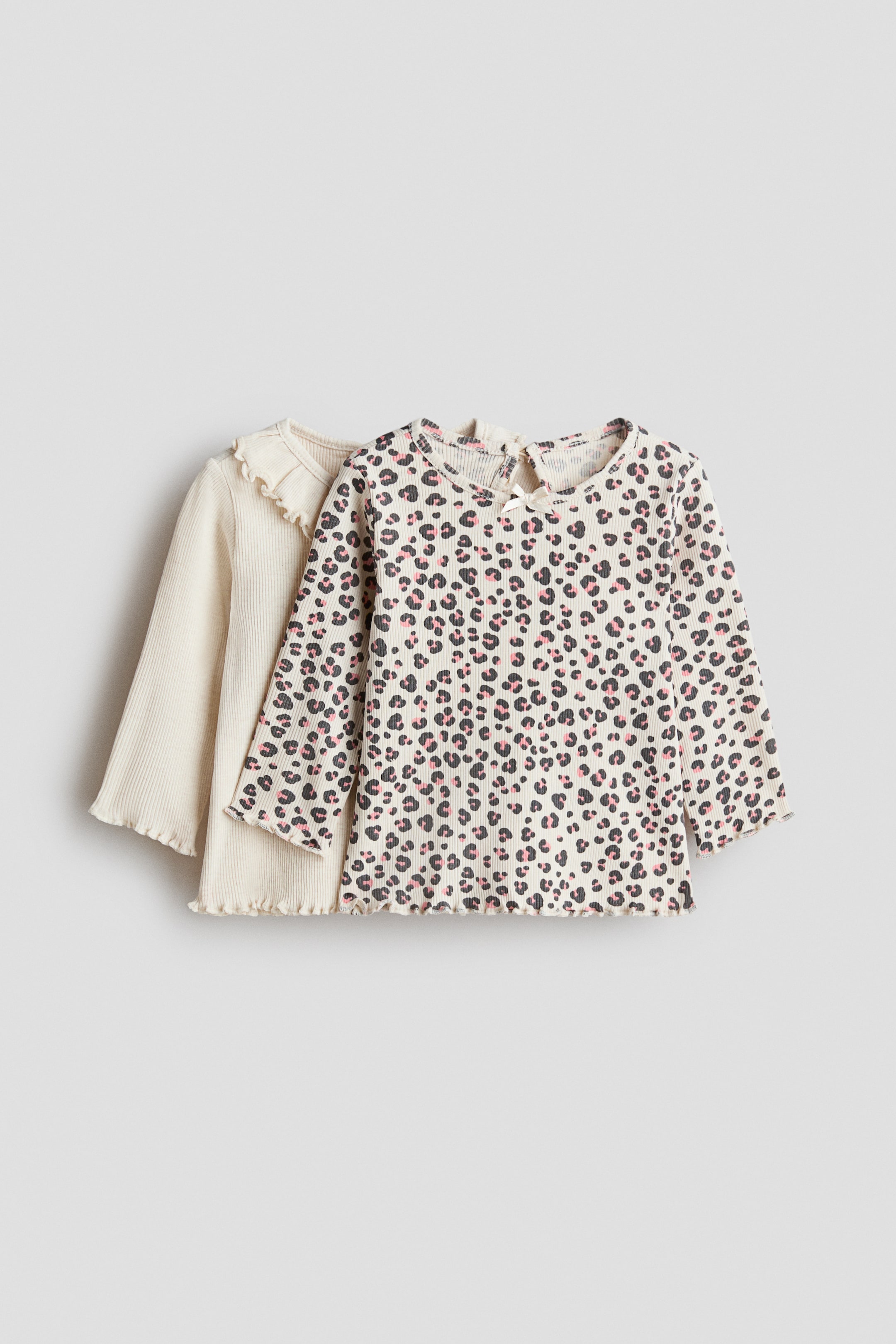 Agrandir l'image: Deux hauts côtelés à manches longues sont présentés, le haut avant comportant une base crème avec un imprimé léopard noir et rose, un col rond, un petit nœud blanc sur la fente au dos, et des manches et un ourlet à volants. Le haut en couches derrière est d'une couleur crème unie, présentant un col à volants, des manches longues, et des manches et un ourlet à volants.
