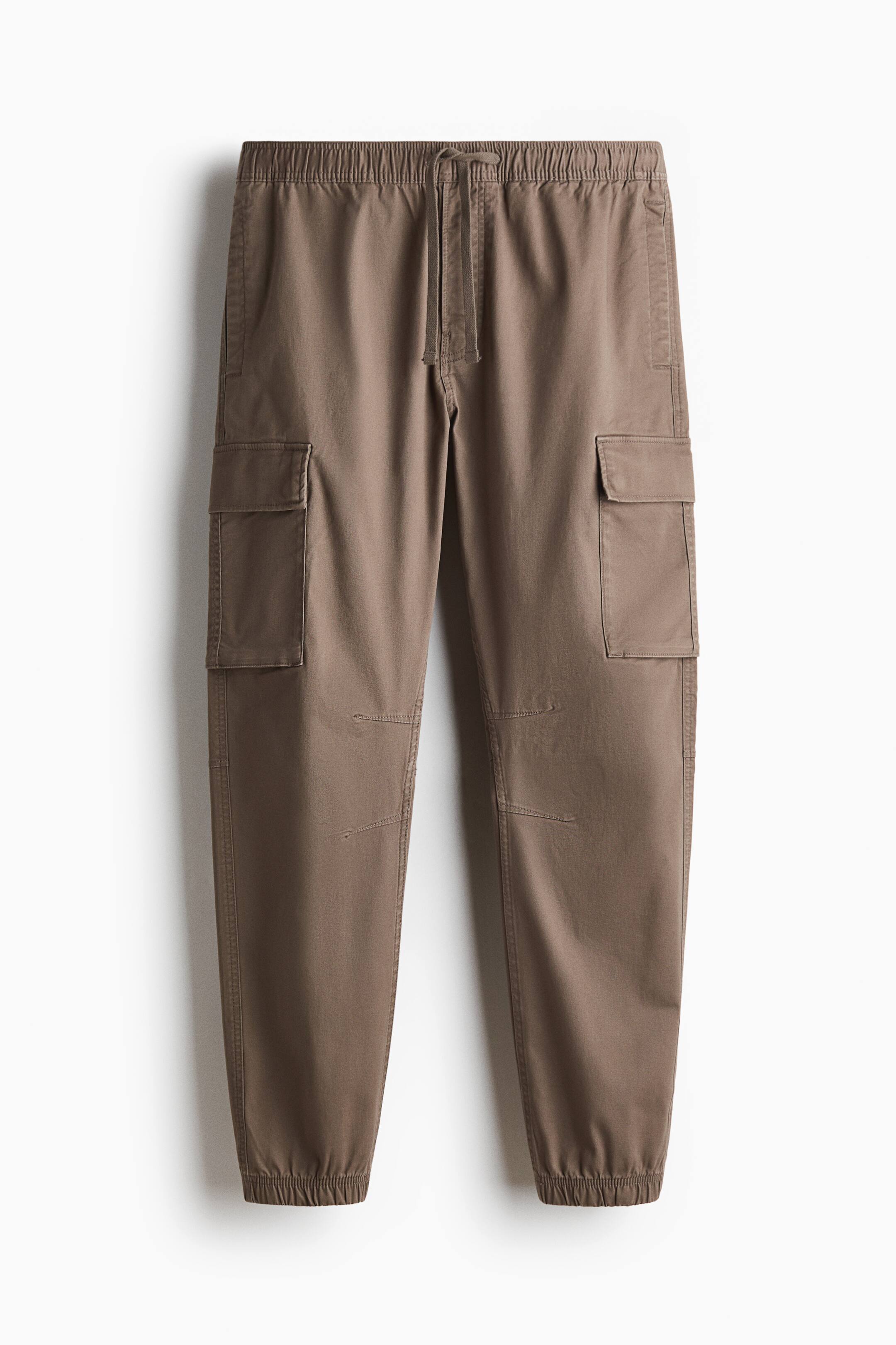 Xem ảnh lớn hơn: Quần dài vải twill cotton màu nâu được trình bày ở mặt trước, với cạp thun có dây rút che kín, khóa giả và túi xéo ở hai bên. Túi cargo nắp được bố trí trên ống quần, cùng ống quần ôm dần, ly gối và gấu bo thun hoàn thiện phom dáng ôm gọn.