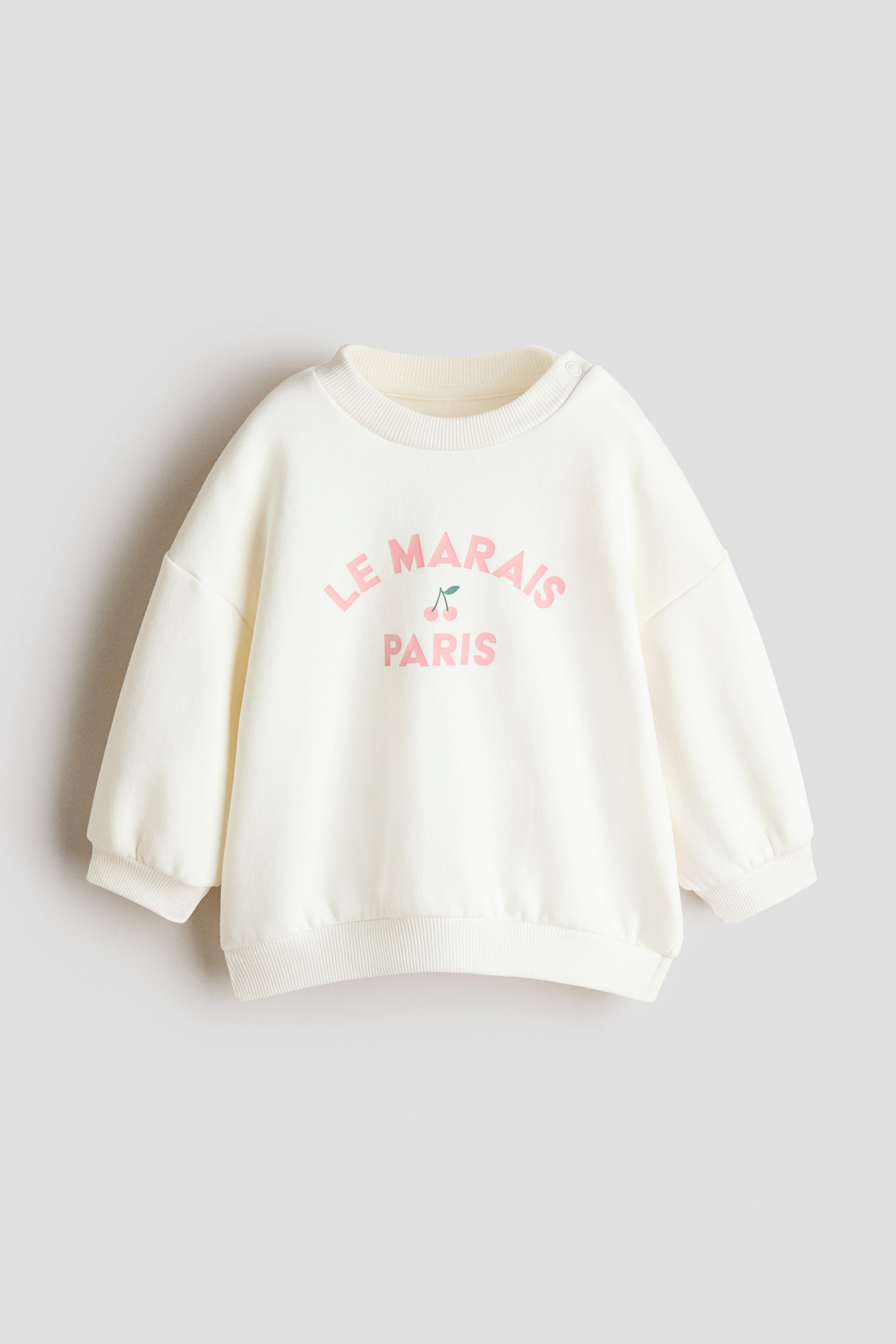 View larger image: A cream sweatshirt is showcasing its front, featuring a ribbed crew neck with two snap buttons on one shoulder. The front displays "LE MARAIS PARIS" in pink lettering accompanied by two small pink cherries, and it is completed with long sleeves, ribbed cuffs, and a ribbed hem.