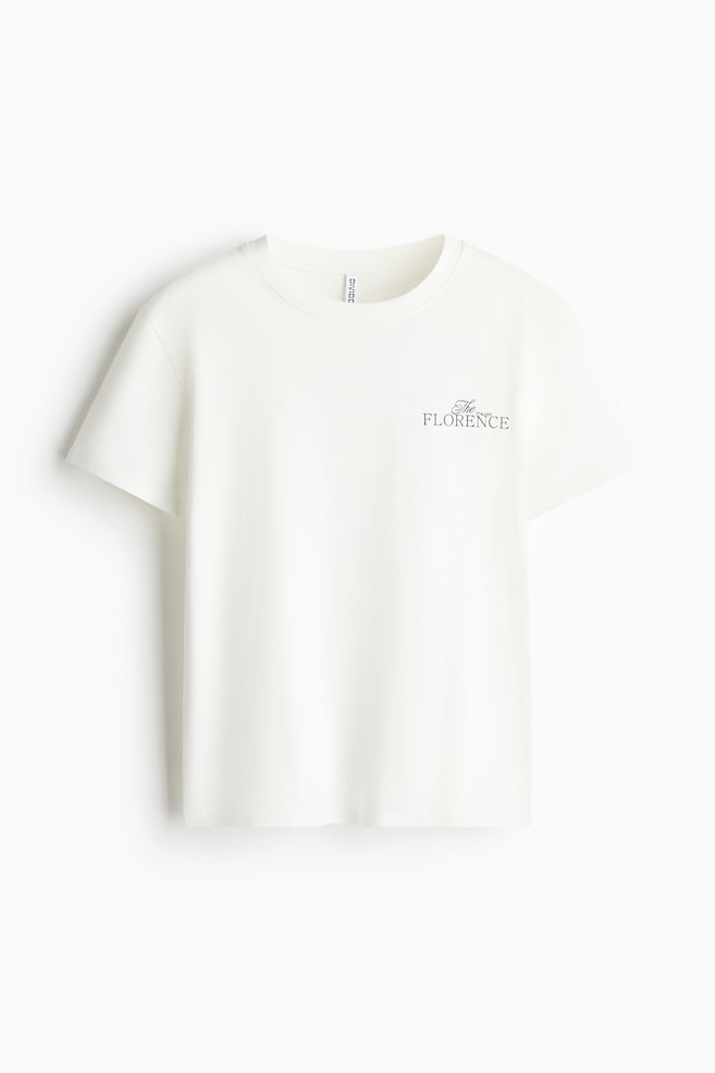 A loose-fit cotton jersey t-shirt in off-white is shown facing front, featuring a ribbed crew neckline, dropped shoulders, and a small black printed text design on the left chest reading "The FLORENCE". - 1