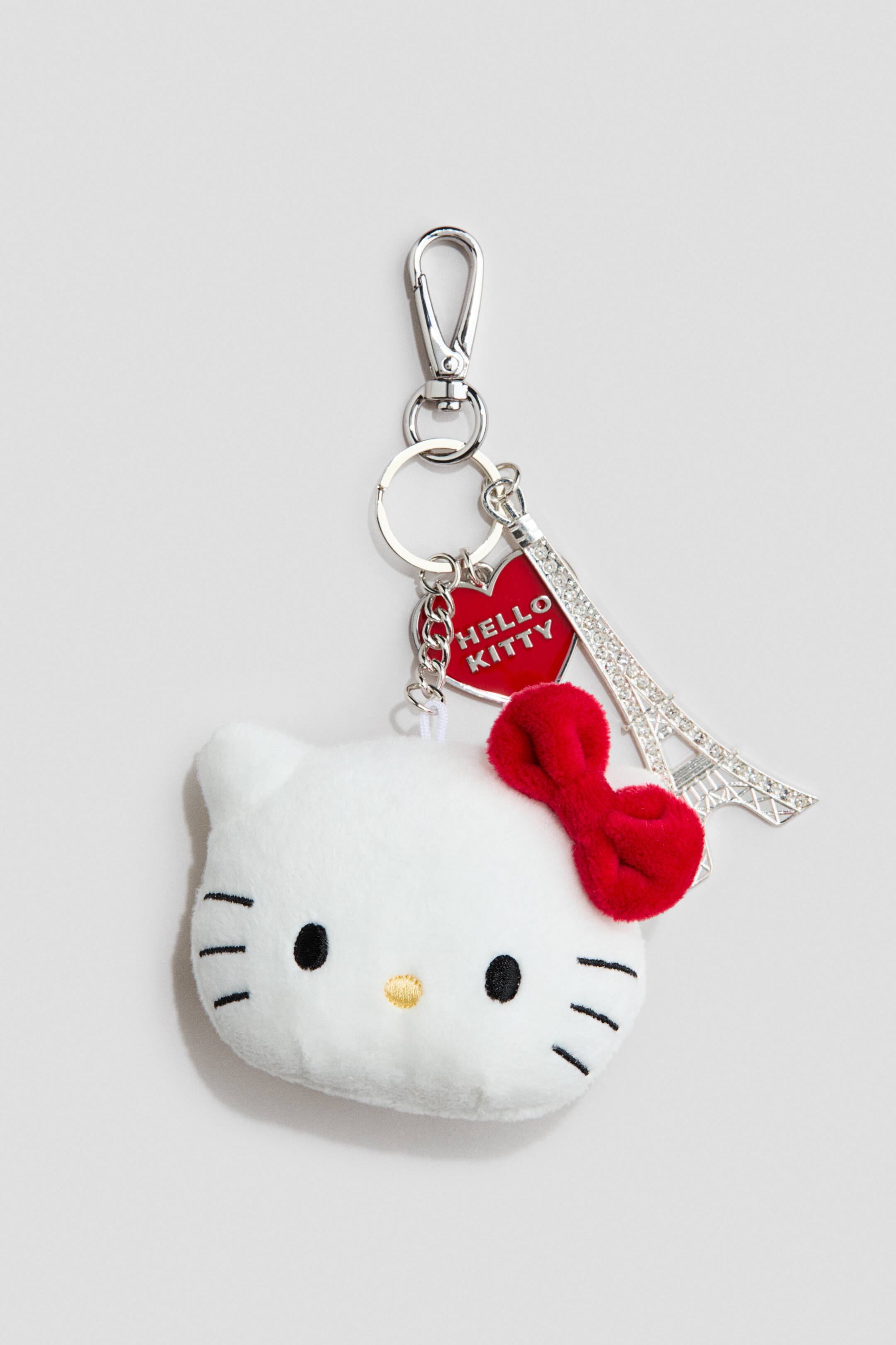 Ampliar la imagen: Un charm para bolso presenta un mosquetón de metal plateado, una anilla partida, un charm de corazón rojo que muestra el texto 'HELLO KITTY', y un charm de la Torre Eiffel adornado con diamantes de imitación. También colgando de la anilla partida hay un charm de cabeza de Hello Kitty de peluche blanco, que muestra ojos ovalados negros, una pequeña nariz ovalada amarilla, bigotes negros y un lazo de peluche rojo en su oreja derecha.
