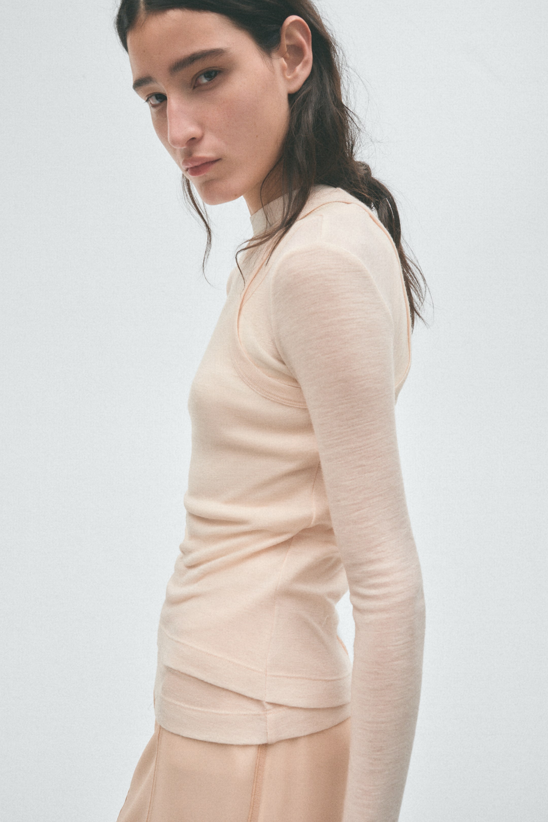 View larger image: The model is wearing a pale peach mock neck long-sleeved top, layered with a matching sleeveless overlay top featuring visible seams.