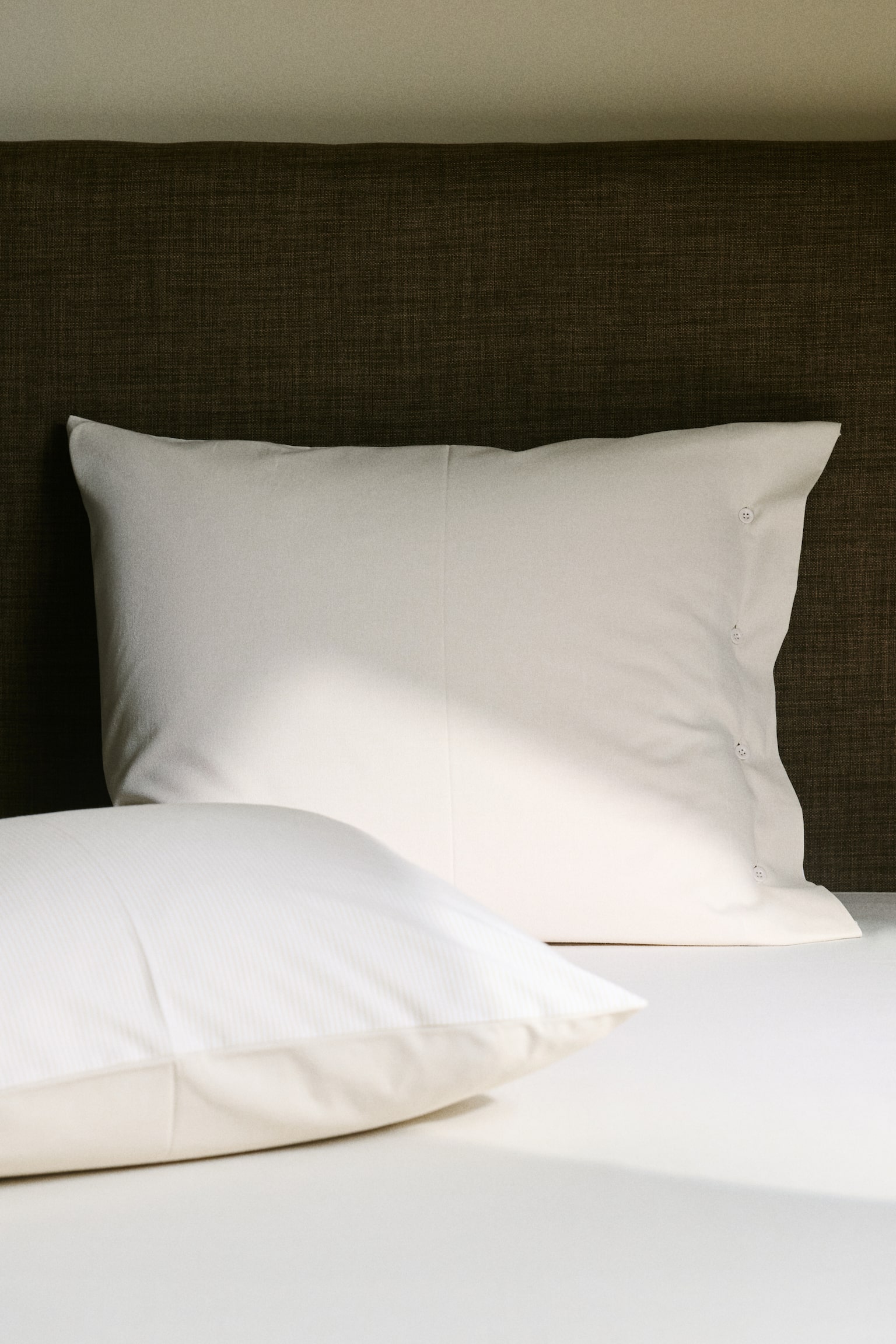 Two off-white rectangular pillows are being presented, each displaying a row of small, round buttons along one vertical edge. The fabric is showing a natural, softly rumpled texture. - 1