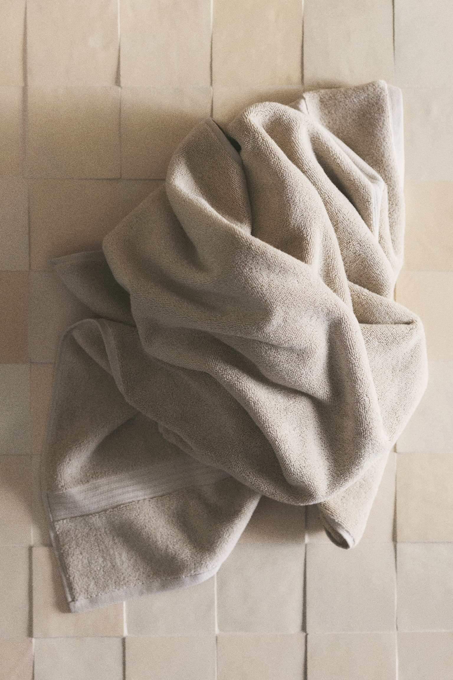 A light beige towel is hanging, folded, showcasing its soft, textured surface. A smoother, horizontal band is visible across the lower section of the visible top fold, with its edges neatly hemmed. - 1