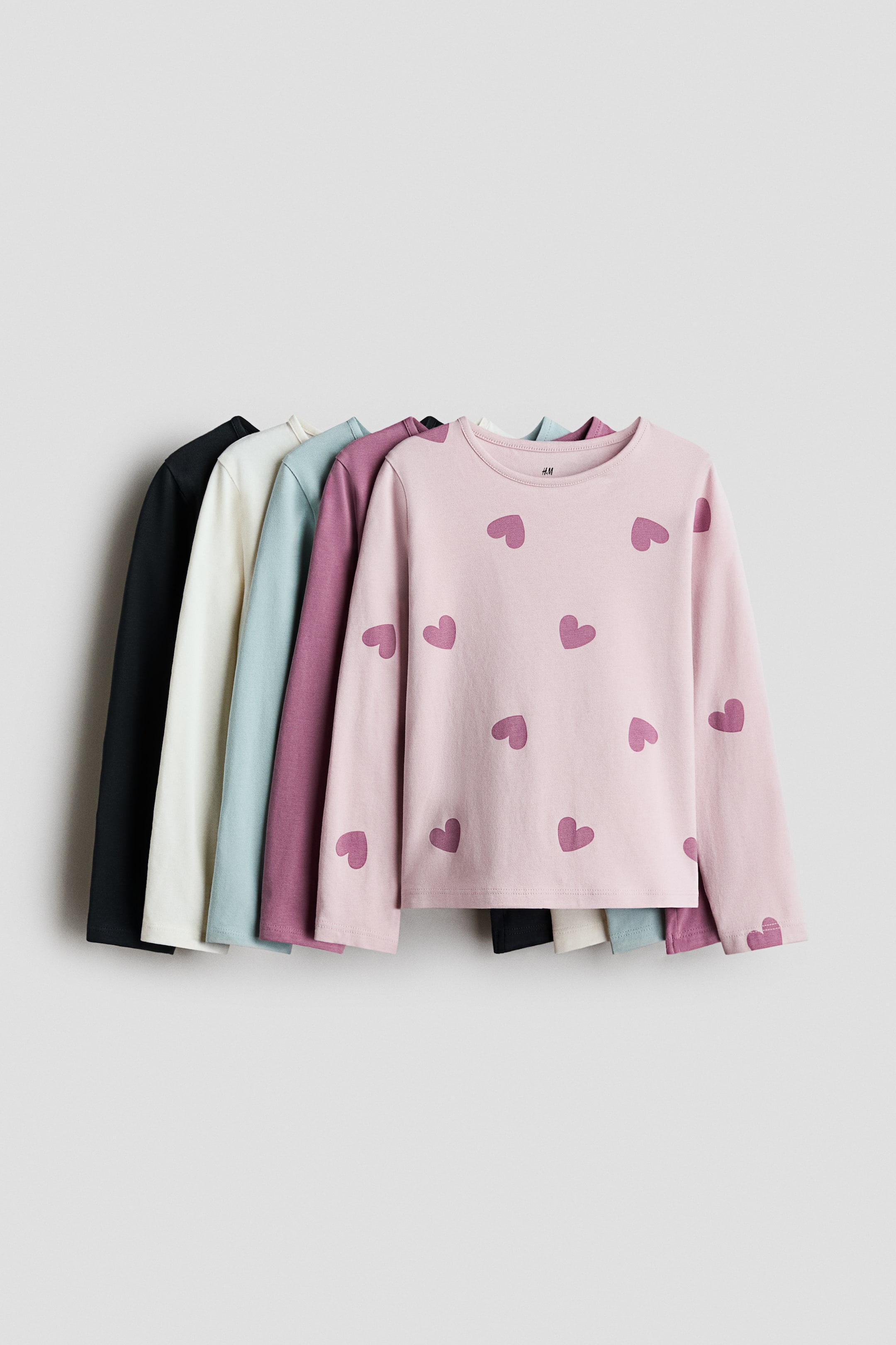 View larger image: Five long-sleeved t-shirts are showcased in a row, all facing front. They feature round necklines and come in black, off-white, light blue, purple, and light pink, with the pink t-shirt adorned with scattered heart motifs.
