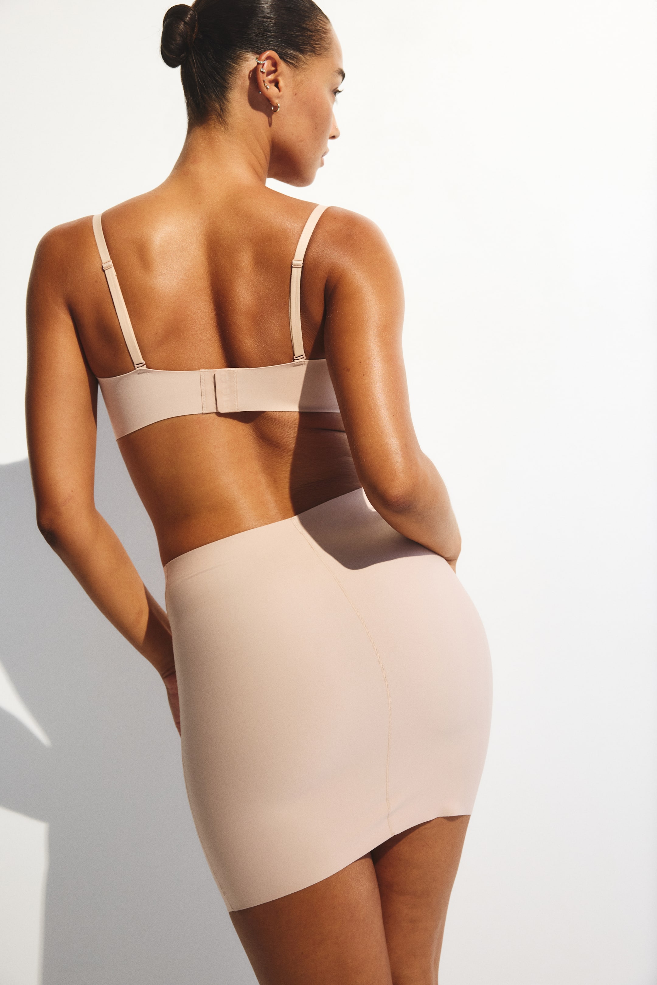 View larger image: A light beige bra and a matching light beige shapewear slip are being presented from the back and a slight side angle. The bra is featuring thin adjustable straps and a hook-and-eye closure, while the seamless slip is smoothly contouring the body and ending above the knee.