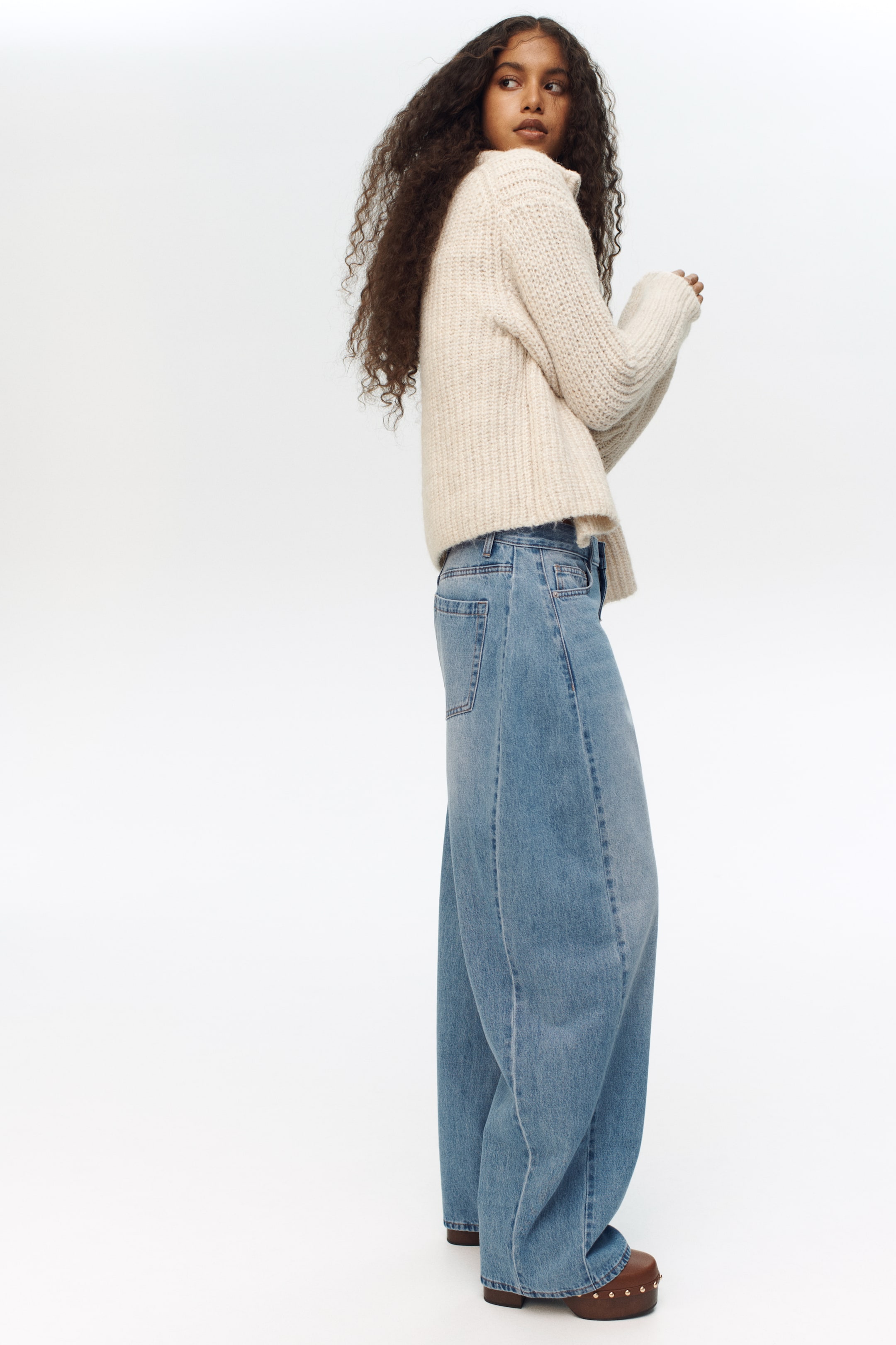 Größeres Bild ansehen: Ein cremefarbener, grob gestrickter Pullover zeigt einen hohen gerippten Kragen und lange gerippte Bündchen. Er ist über einer hell gewaschenen, weit geschnittenen Jeans mit auffälligen vorderen Nähten und einer sichtbaren Gesäßtasche auf der linken Seite geschichtet.