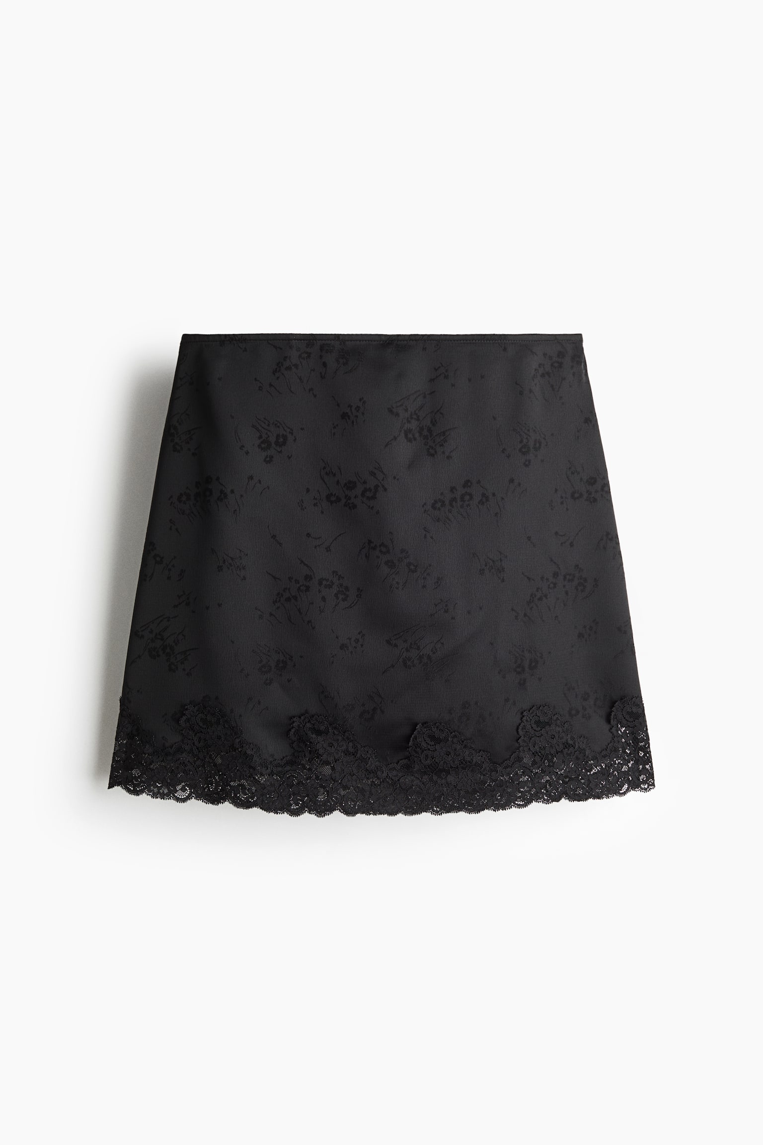 A black mini skirt is shown from the front, featuring a subtle tonal floral jacquard pattern throughout the fabric. The design is finished with a wide scalloped black lace trim along the bottom hem.
