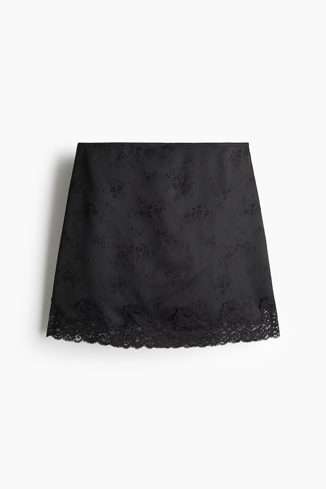 A black mini skirt is shown from the front, featuring a subtle tonal floral jacquard pattern throughout the fabric. The design is finished with a wide scalloped black lace trim along the bottom hem. - 1