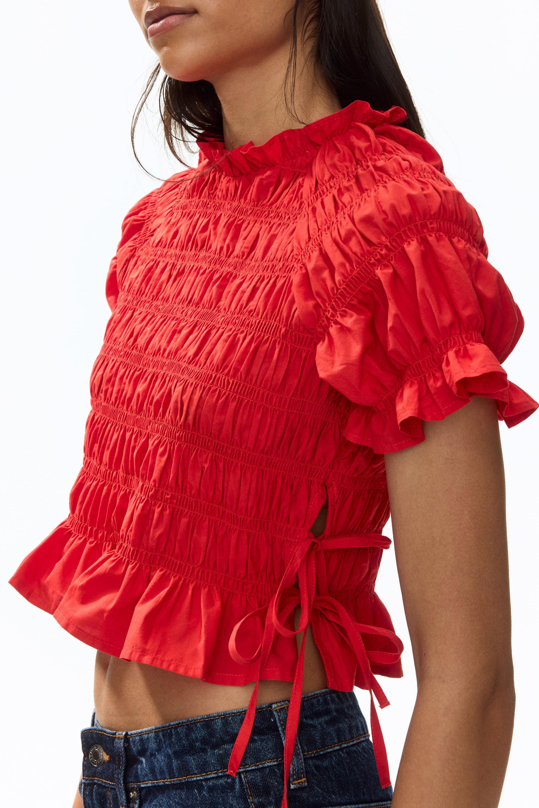 View larger image: The model is donning a short, fitted red smocked blouse. It boasts frill trims along the round neckline and short raglan sleeves, featuring a side slit on the hem with ties that are elegantly fastened.